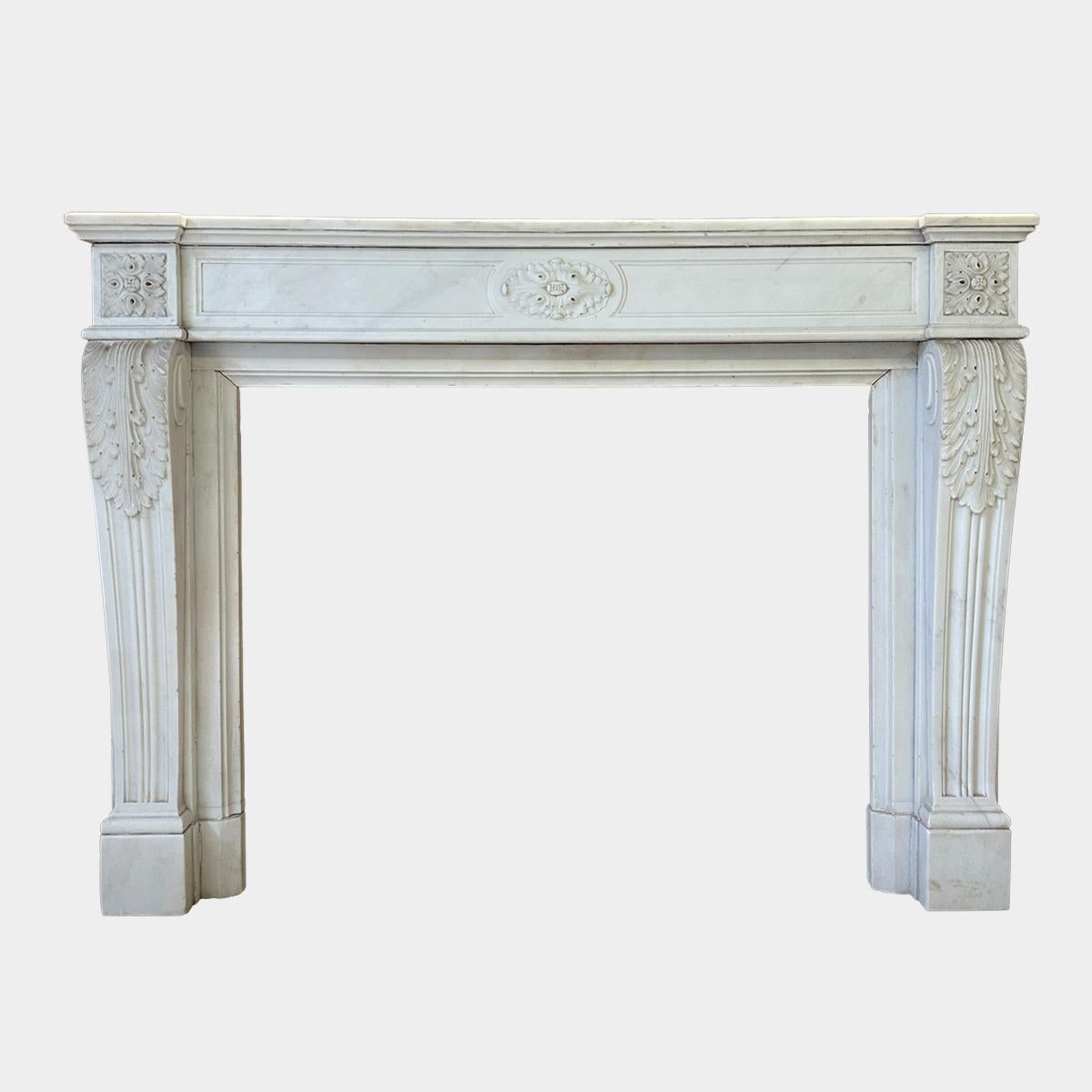 Cheminée en marbre blanc statuaire Louis XVI de la fin du XVIIIe siècle. Les montants de la console sont effilés et chantournés, les sommets sont finement sculptés de feuilles d'acanthe, et le panneau frontal est à deux champs et cannelé. Le