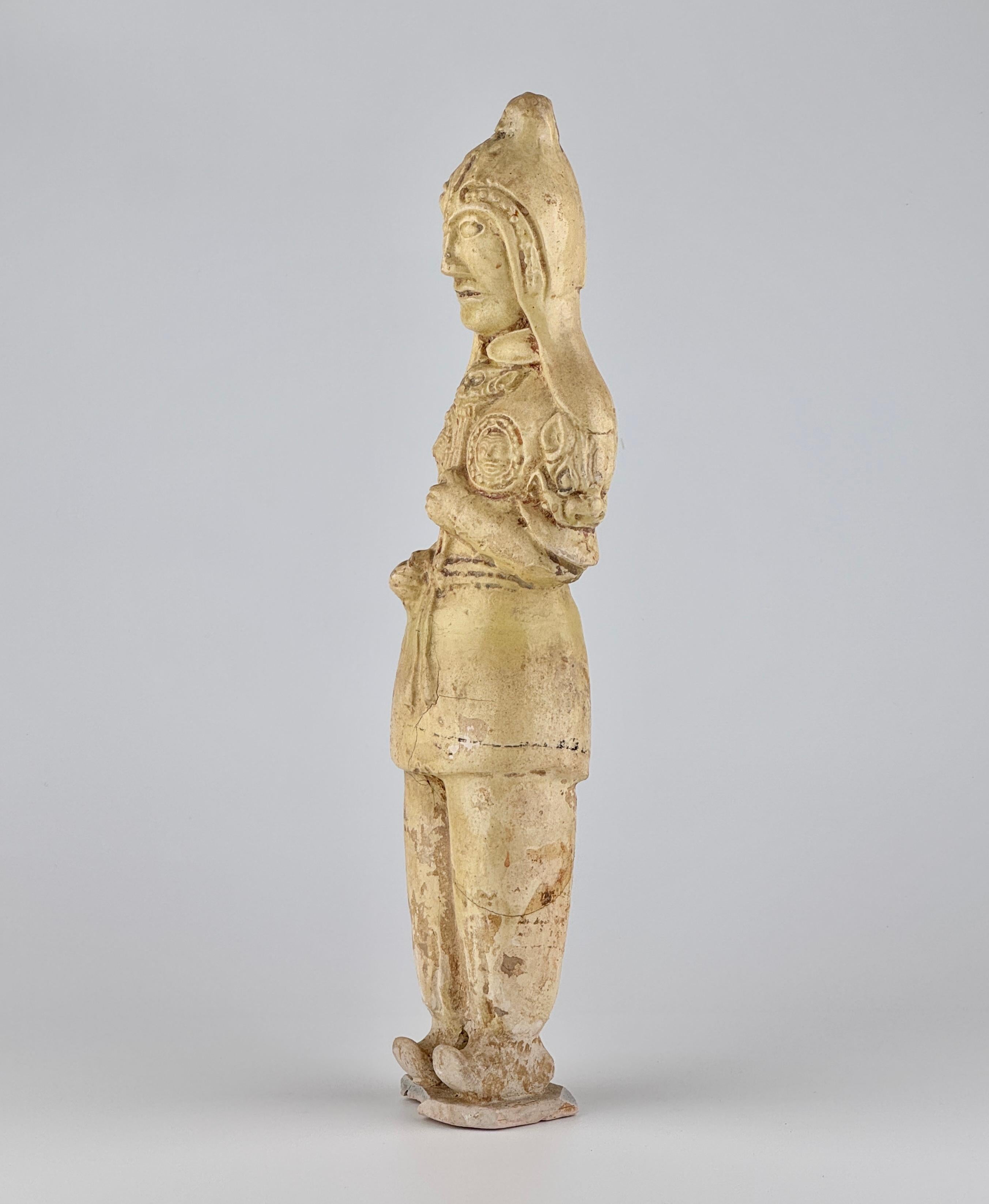This well-modeled ceramic figure depicts a Scythian warrior in full armor, likely serving as a standard bearer. The warrior stands upright on a thin rectangular base, with clenched fists suggesting he once held a standard. His facial features are