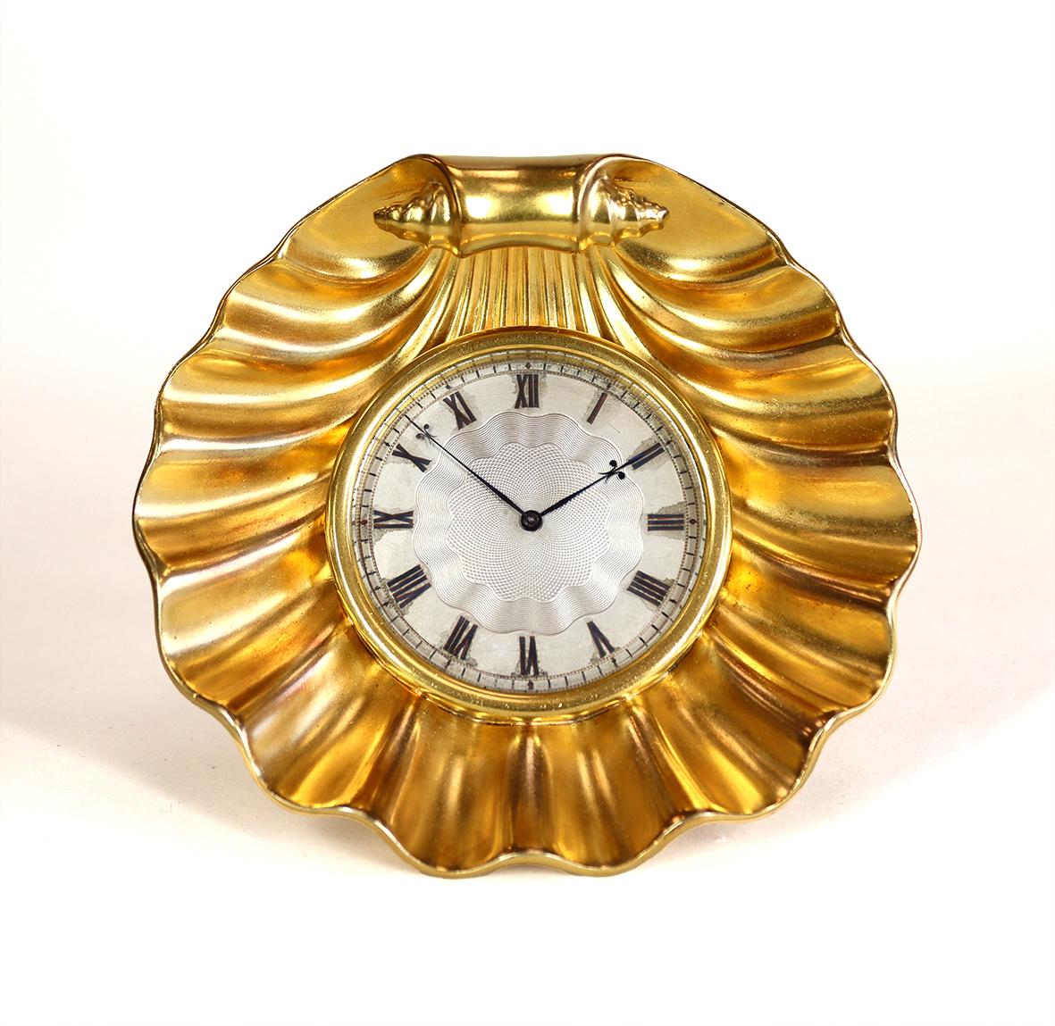 This beautiful desk timepiece is a rare strut clock, in the form of a gilded scallop shell, with an engine turned silvered roman dial set behind a bevelled glass with distinctive fleur de lis hands. The the fine movement has an excellent lever
