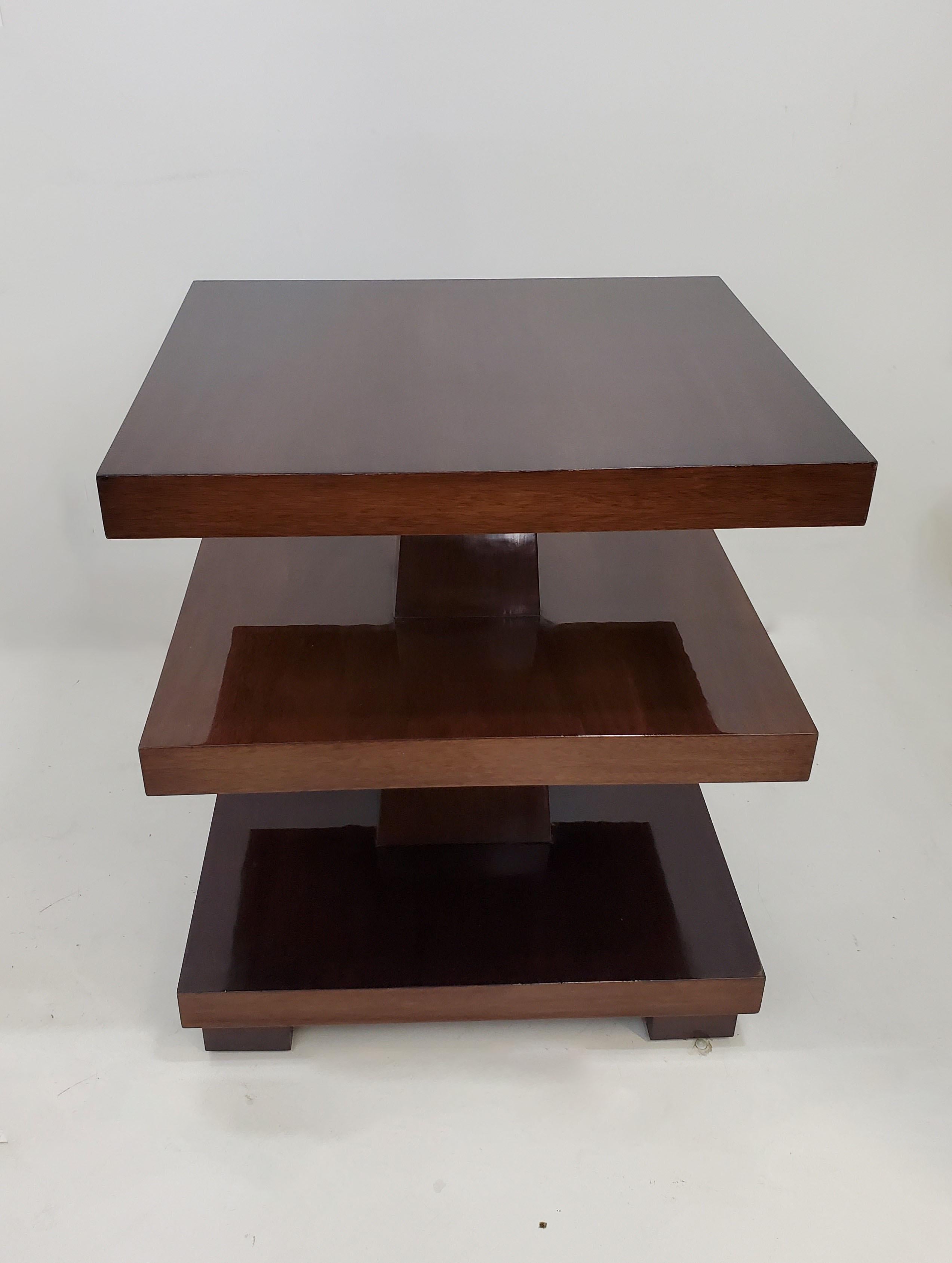 An elegant and imposing highly sculptural, Art Deco revival rectangular three tiered table of cubist design in a deep cognac /dark amber colored mahogany with central shaft dissecting the shelves and the whole raised on cube feet.
The angular form