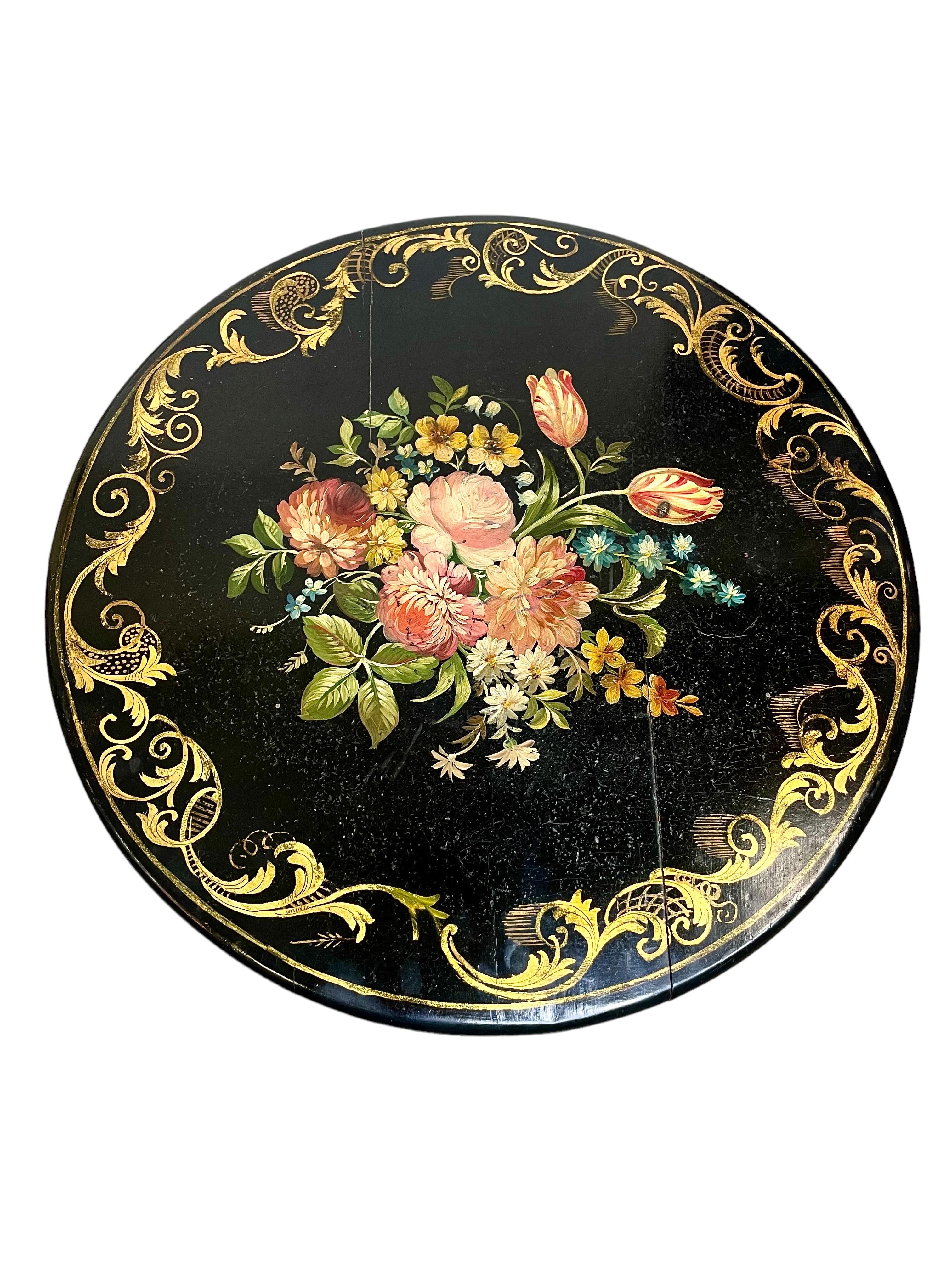 A stunning Napoleon III style ebonised guéridon table, with a lacquered circular top, and featuring a swirling parcel gilt decoration around the perimeter and a hand-painted polychrome design of wild flowers and foliage at the centre. The table is