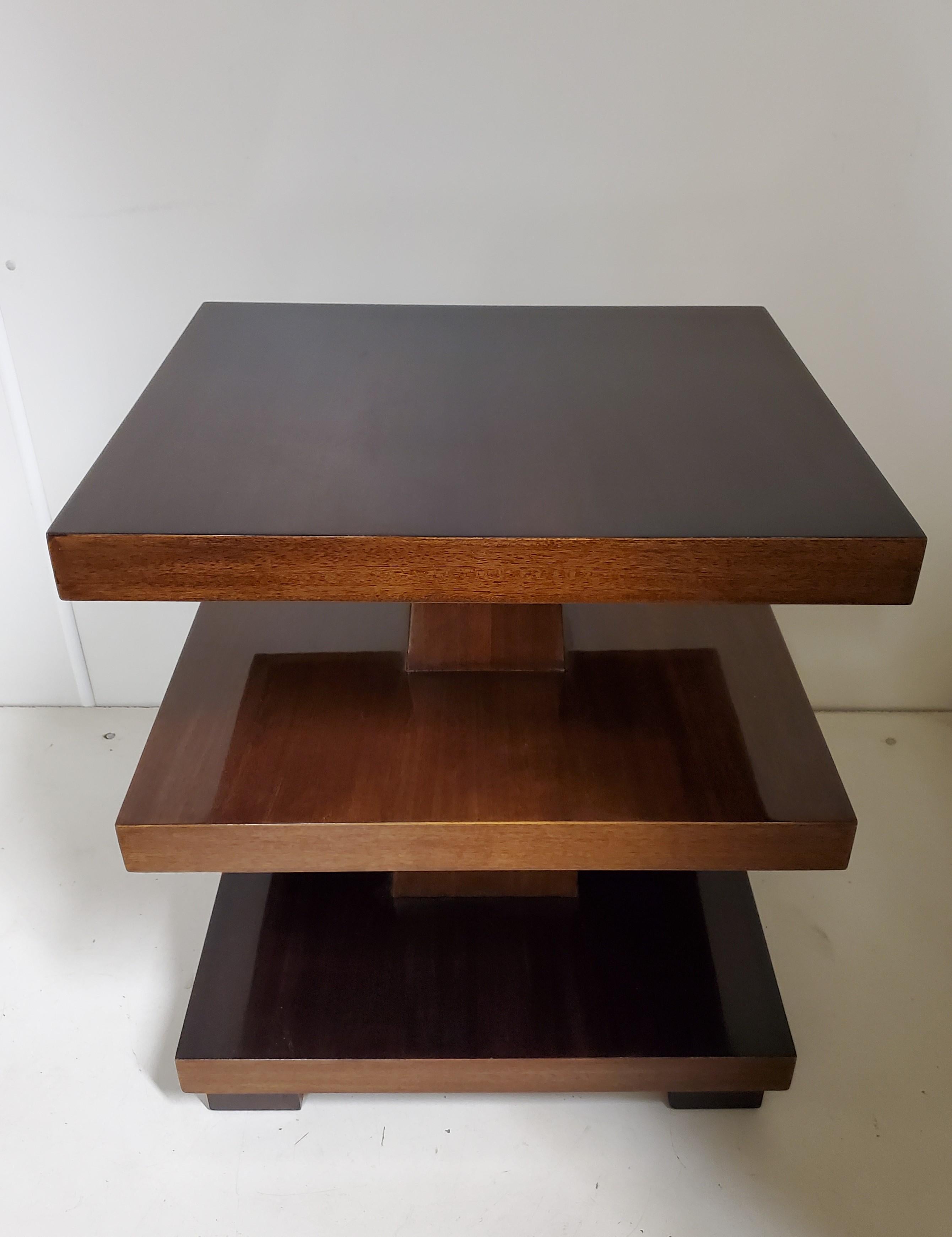 An elegant and imposing highly sculptural, Art Deco rectangular three tiered tables of cubist design in a deep cognac /dark amber colored mahogany with central shaft dissecting the shelves and the whole raised on cube feet.
The angular form