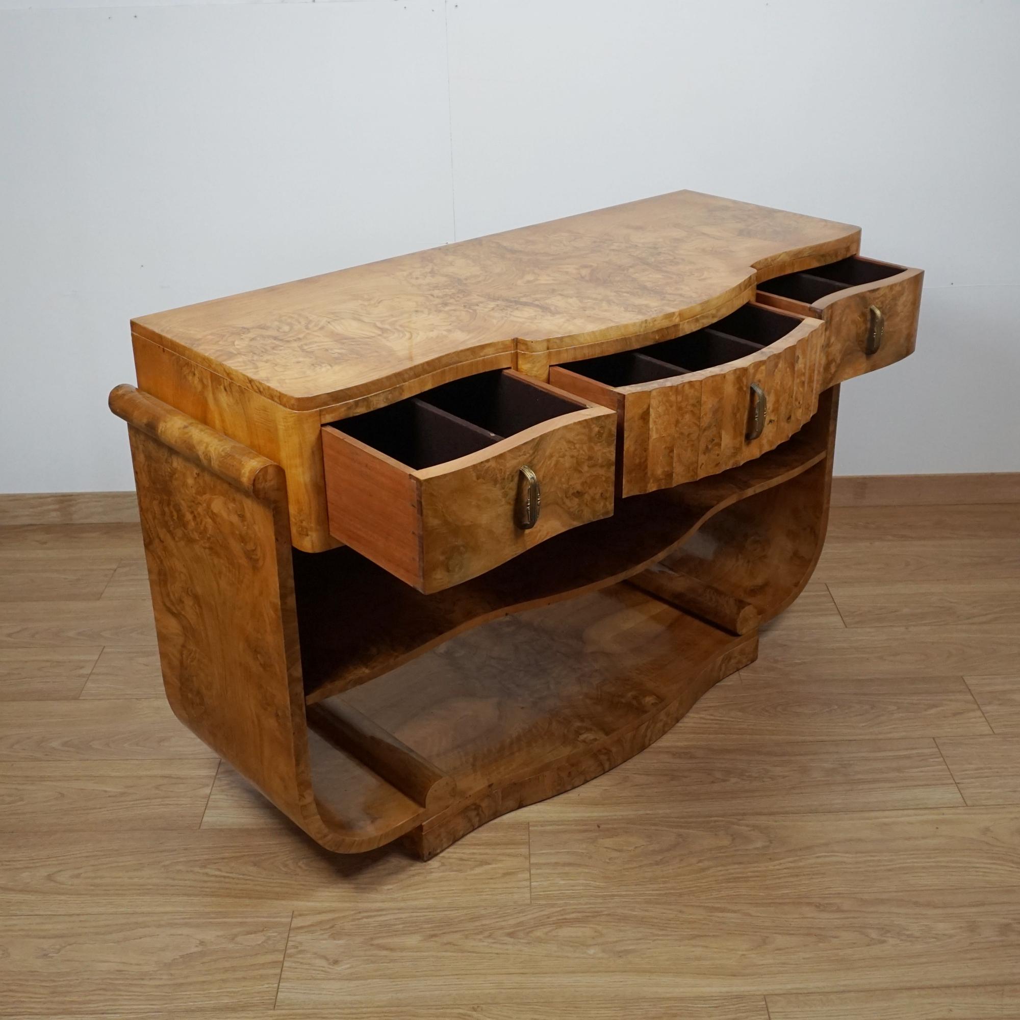 Ein Art-Deco-Konsolentisch von Harry & Lou Epstein. Gratnussbaum furniert auf massivem Mahagoni mit originalen Griffen. Auf einem U-förmigen Sockel mit unterer Ablage und drei oberen Schubladen. 

Abmessungen: H 85cm B 132cm T 52cm

Herkunft: