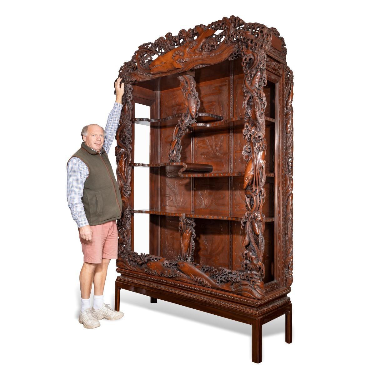 A superb monumental Meiji period hard wood display cabinet, by Noguchi of Yokahama, of rectangular form with three shelves which curl back under themselves like ripples to create an asymmetrical appearance, the ornate frame boldly carved with