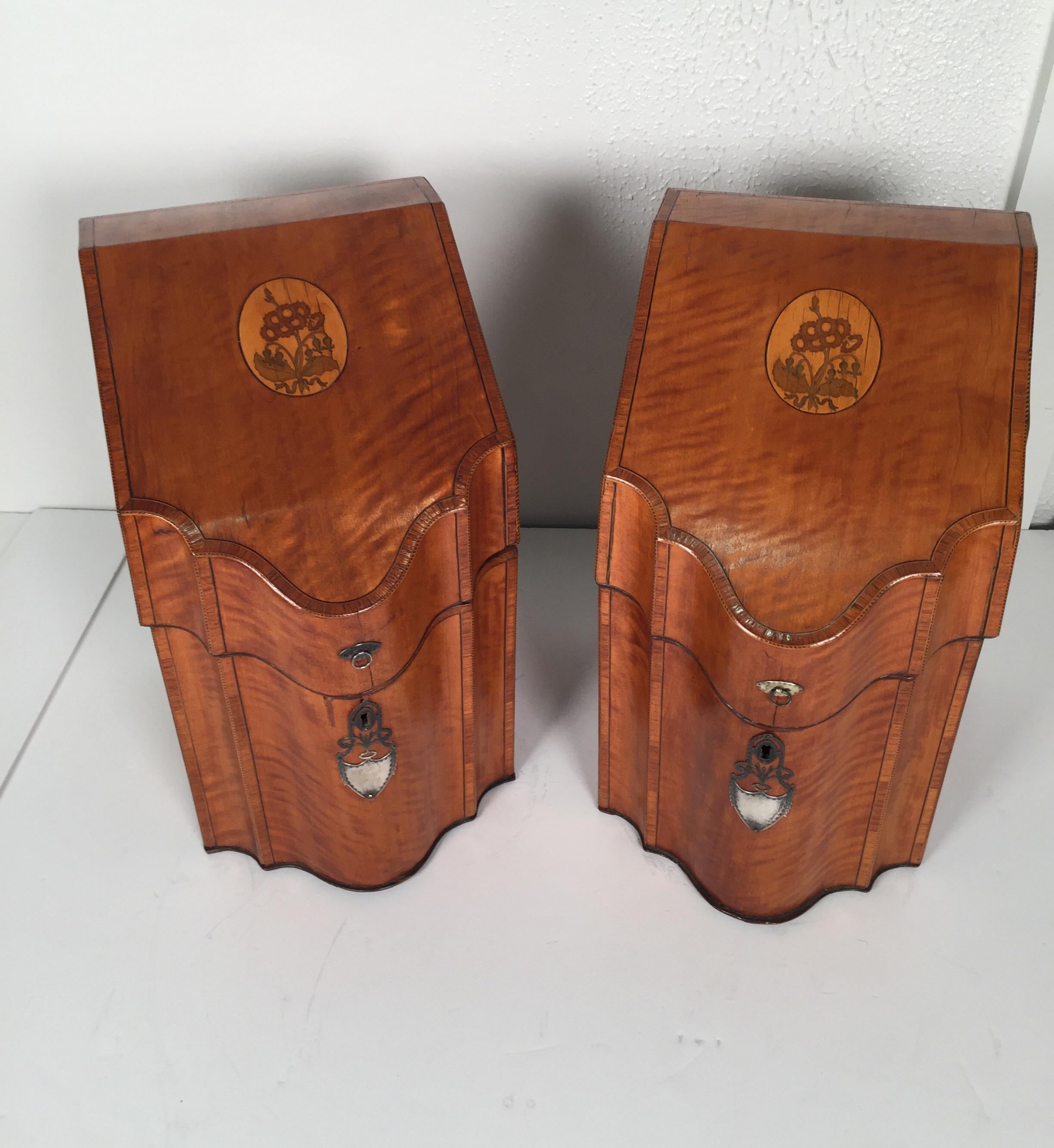 A pair of English satinwood knife boxes with sterling escutcheons. The tops open to reveal the original fittings. The original pair with serpentine fronts and sloped top. The boxes in very good condition with warping from age. No keys.