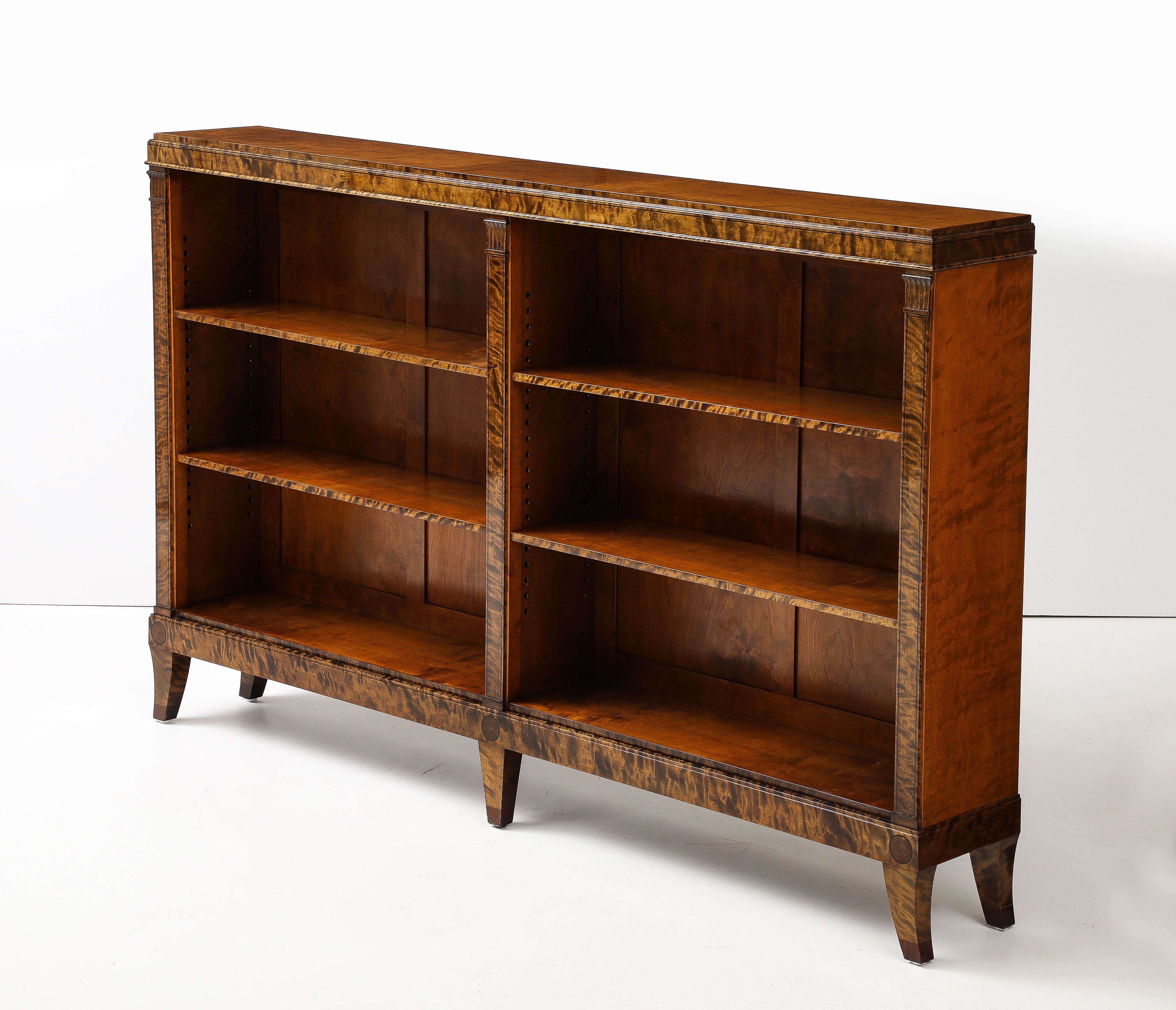 A Swedish Grace flamed birch open bookcase/cabinet, Circa 1930-40, probably designed by Erik Chambert, the stepped rectangular top above two shelves per side, flanked by flat pilasters headed with carved capitals, raised on sabre legs. Adjustable