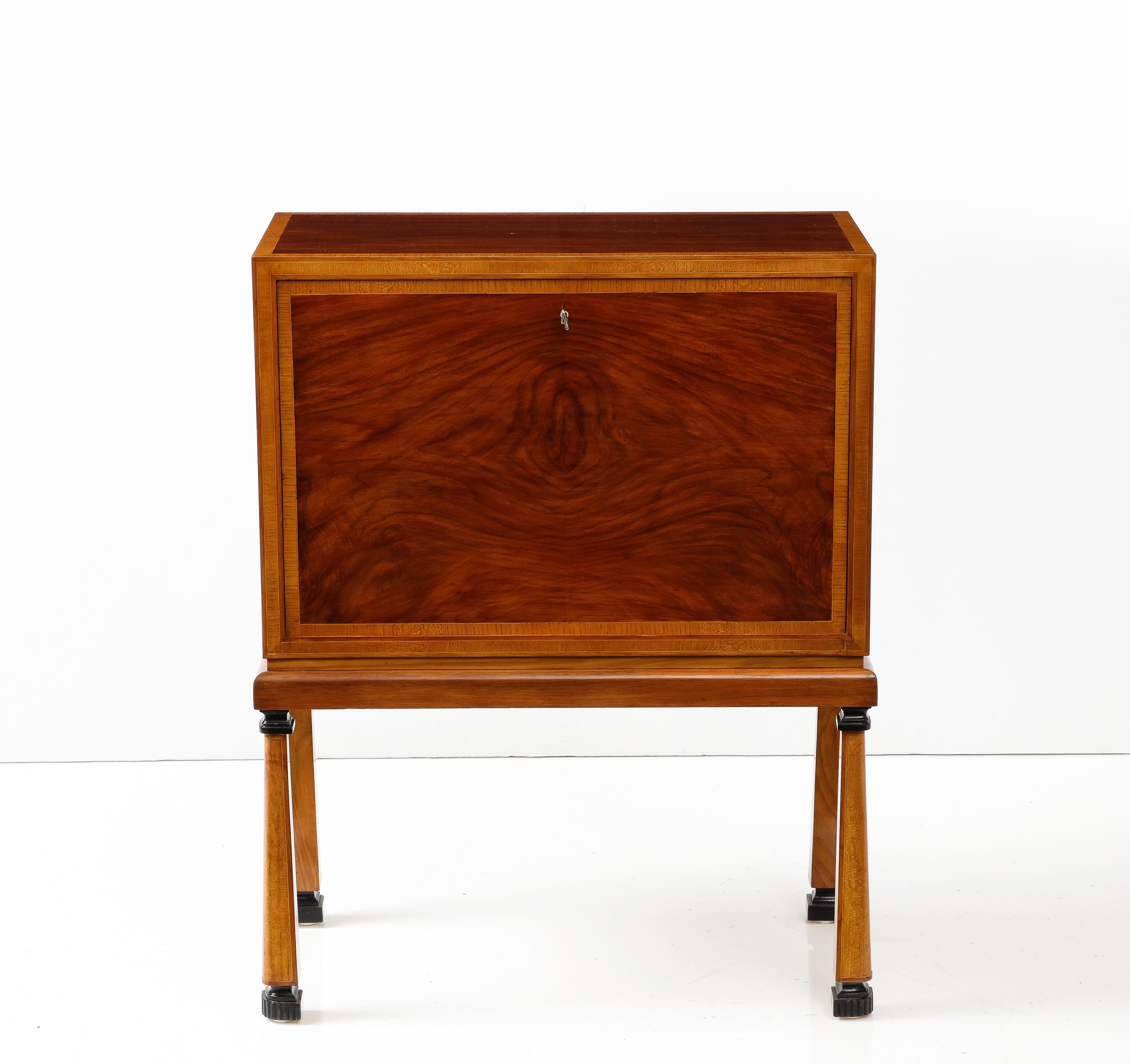 A Swedish Grace nut wood cabinet, circa 1930s, the rectangular top with fruitwood cross-banding above a fall front flap. The interior with four long drawers, raised on square spreading legs headed and ending with ebonized carved moldings. 29.5