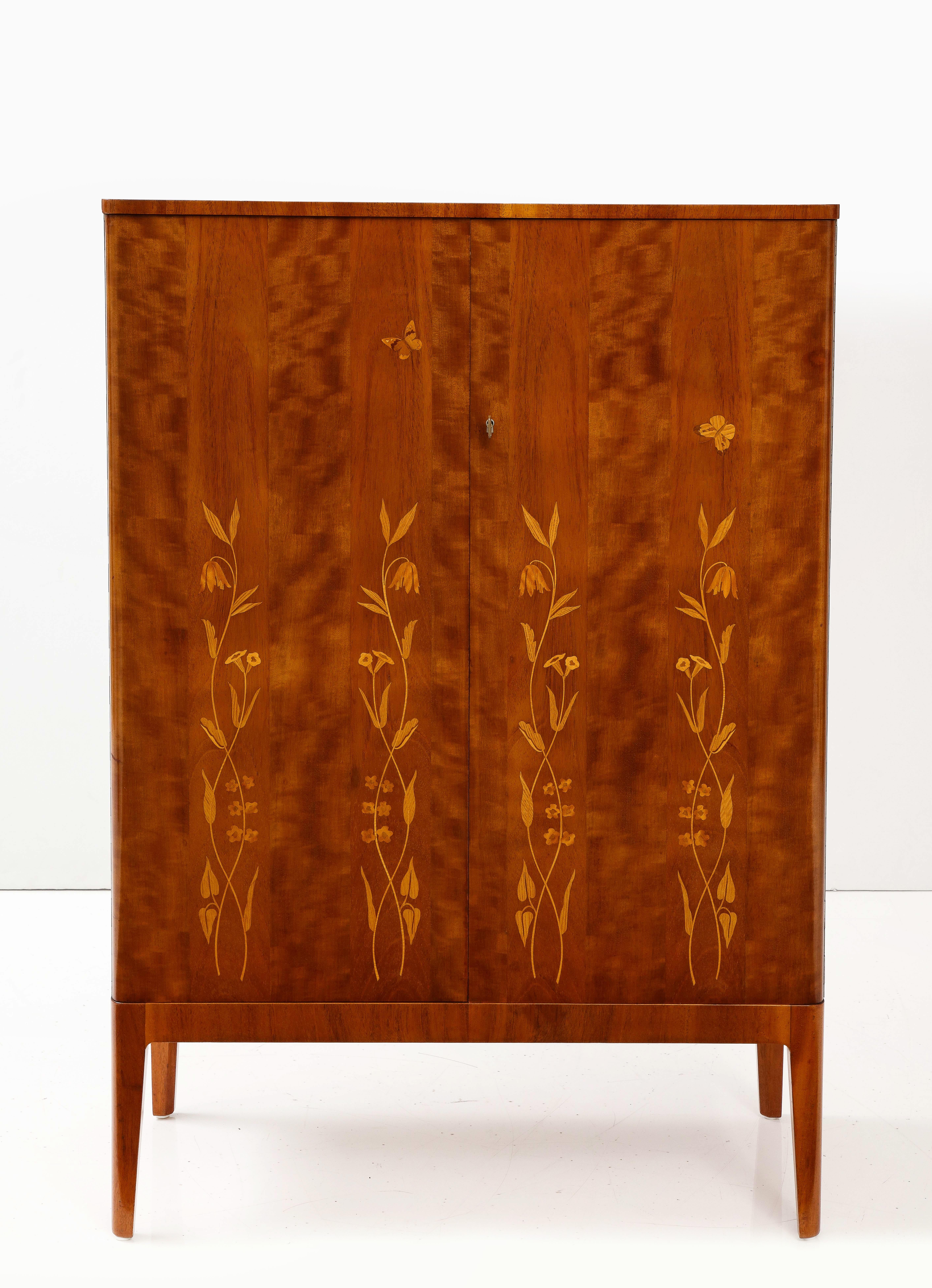 A Swedish Grace mahogany cabinet, Circa 1940s, the rectangular top with slightly rounded forcorners, above two cupboard doors with fruitwood leaf, flower and butterfly inlays, raised on tapered legs. The interior with four drawers above two shelves.