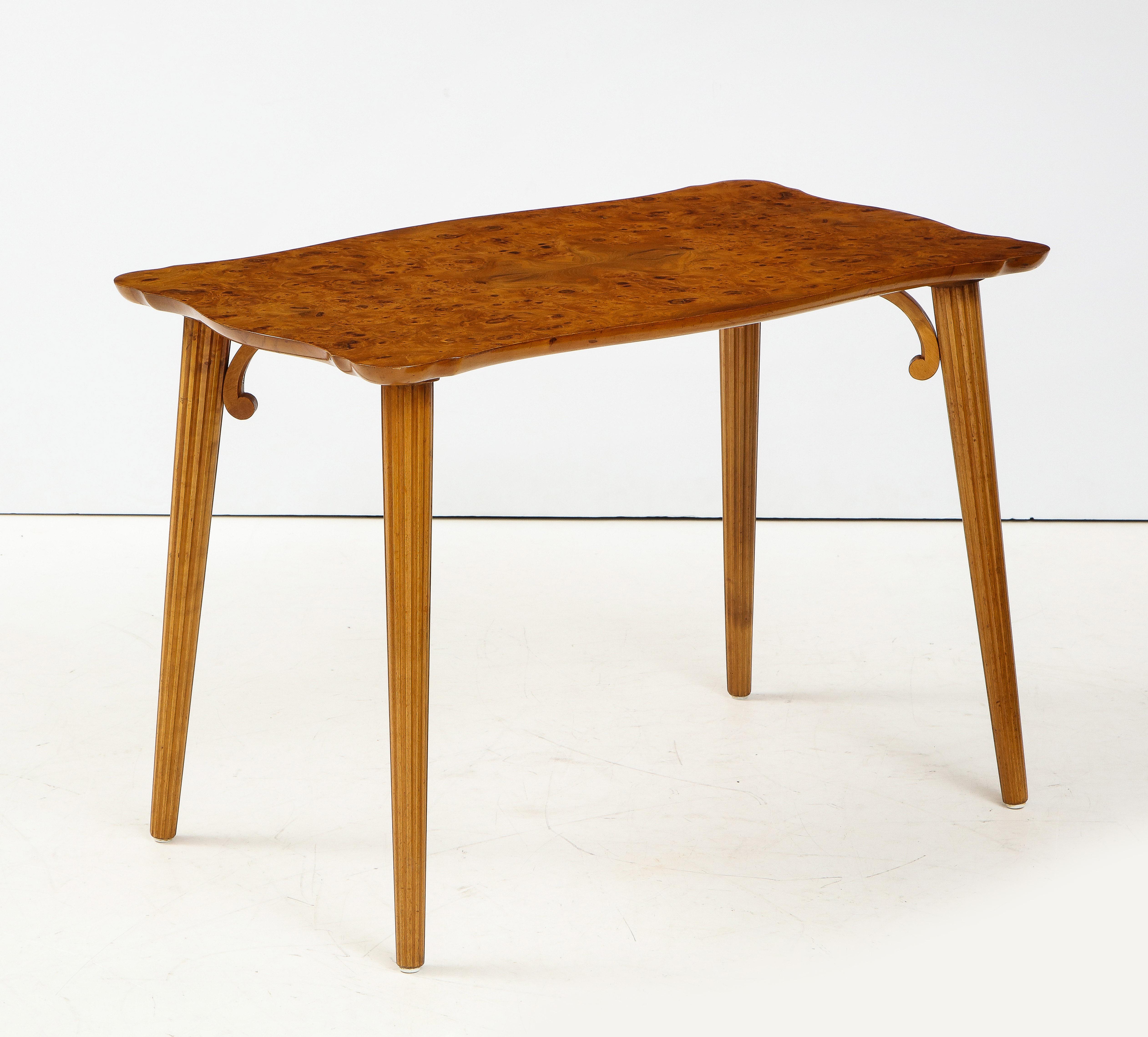 A Swedish Modern burr birch side table, Circa 1940s, the figured burr birch rectangular top with a shaped edge raised on circular tapered and fluted legs headed with a scrolled support.