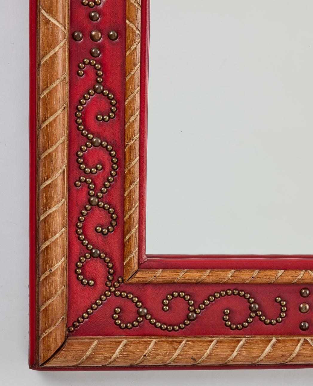 Uno specchio moderno svedese con una cornice in Oak avvolta in pelle artificiale rosso intenso. La cornice è impreziosita da chiodi in ottone perlato con motivi a volute ornamentali. I dettagli intagliati diagonalmente in Oak accentuano i bordi,