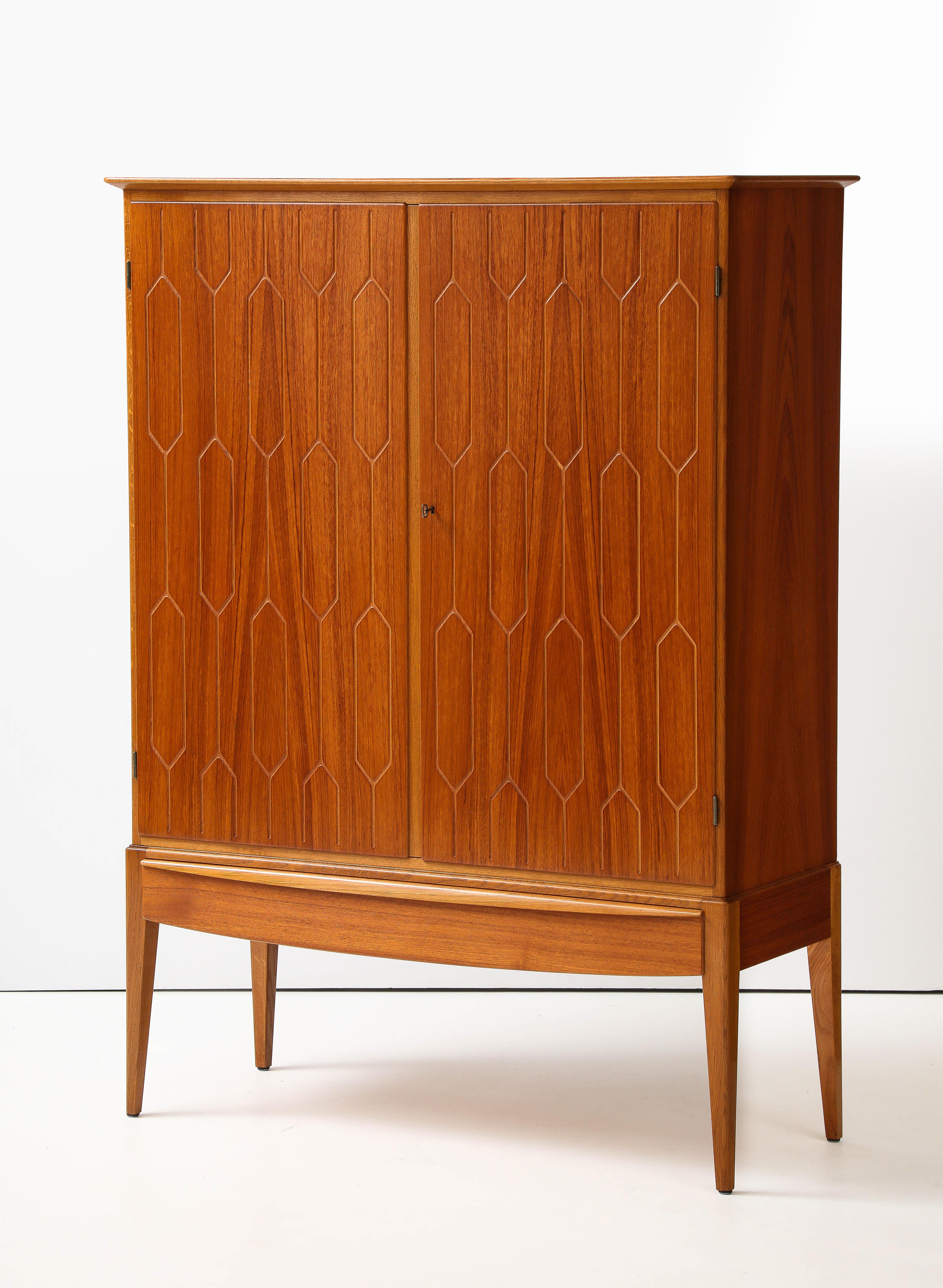 Armoire suédoise moderniste en teck, Circa 1940-1950, le dessus rectangulaire en surplomb au-dessus de deux portes de placard avec un motif hexagonal incisé, au-dessus d'un long tiroir. Monté sur des pieds carrés effilés. Etagères réglables, deux