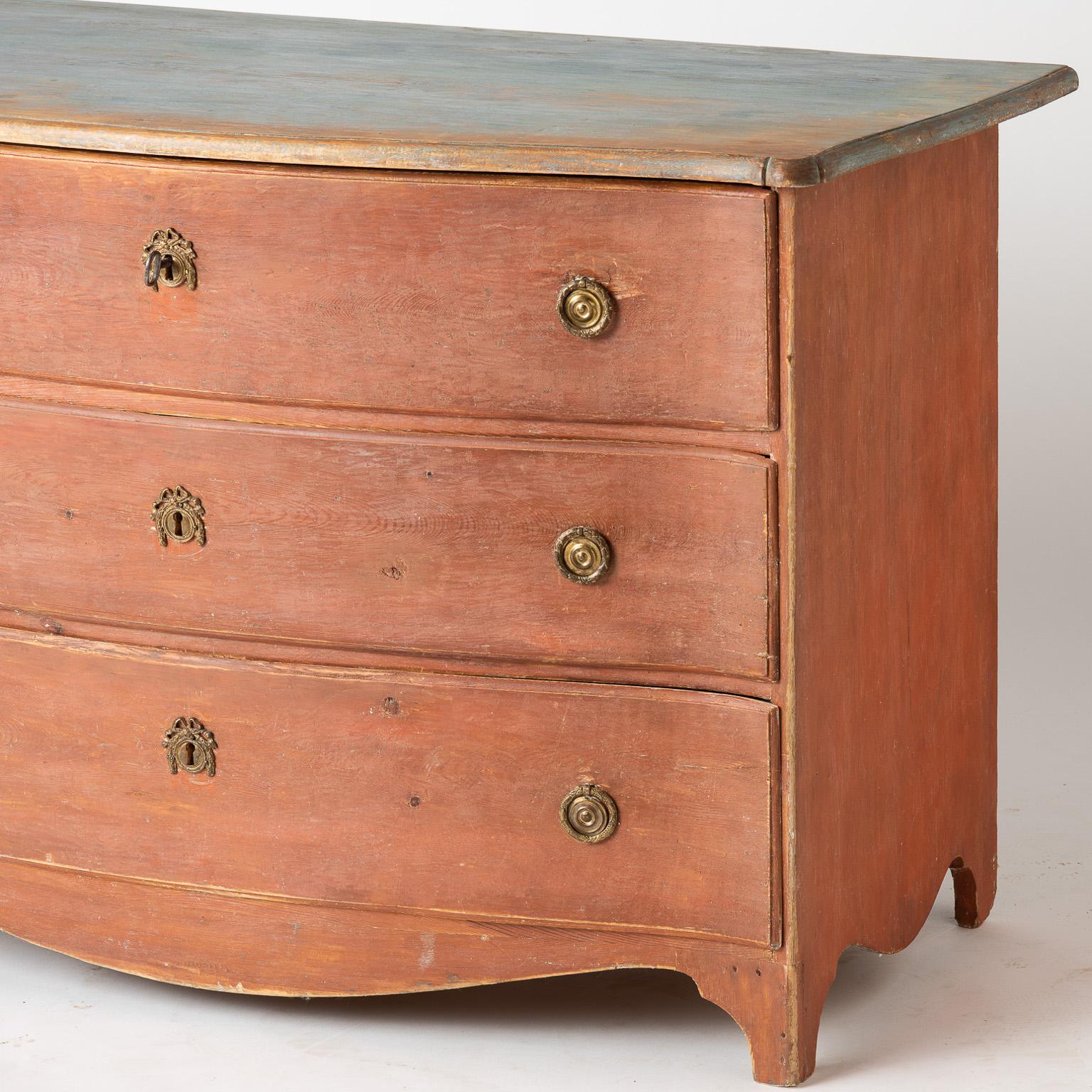 This graceful serpentine chest, in a wonderful shade of coral paint, comes from the north of Sweden. It has the original hardware featuring a brass bow detail on the drawer pulls and escutcheons. The traces of original blue paint on the top have