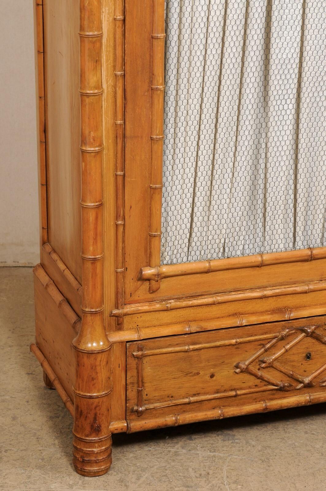 A tall English cabinet with faux bamboo design and glass panel front door, from the early 20th century. This antique curio cabinet from England features a beautiful raised pediment top with finials adorning the front side posts. It has been