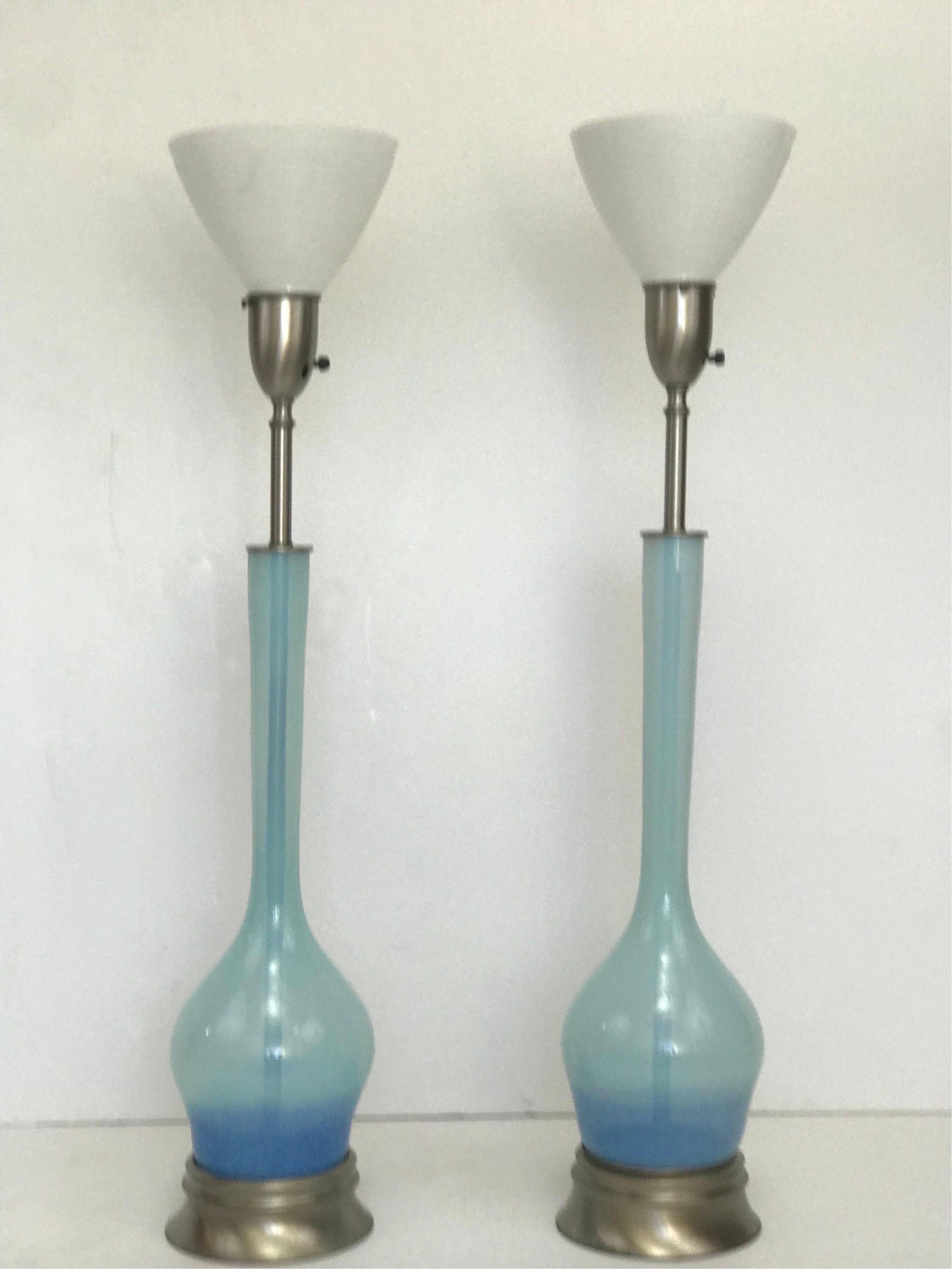 A tall pair of early 1950’s transitional modern table lamps in hand blown blue glass that is slightly graded from top to bottom. Both are slightly different than each other yet a matched pair and transition from an opaque silvery blue at the neck to