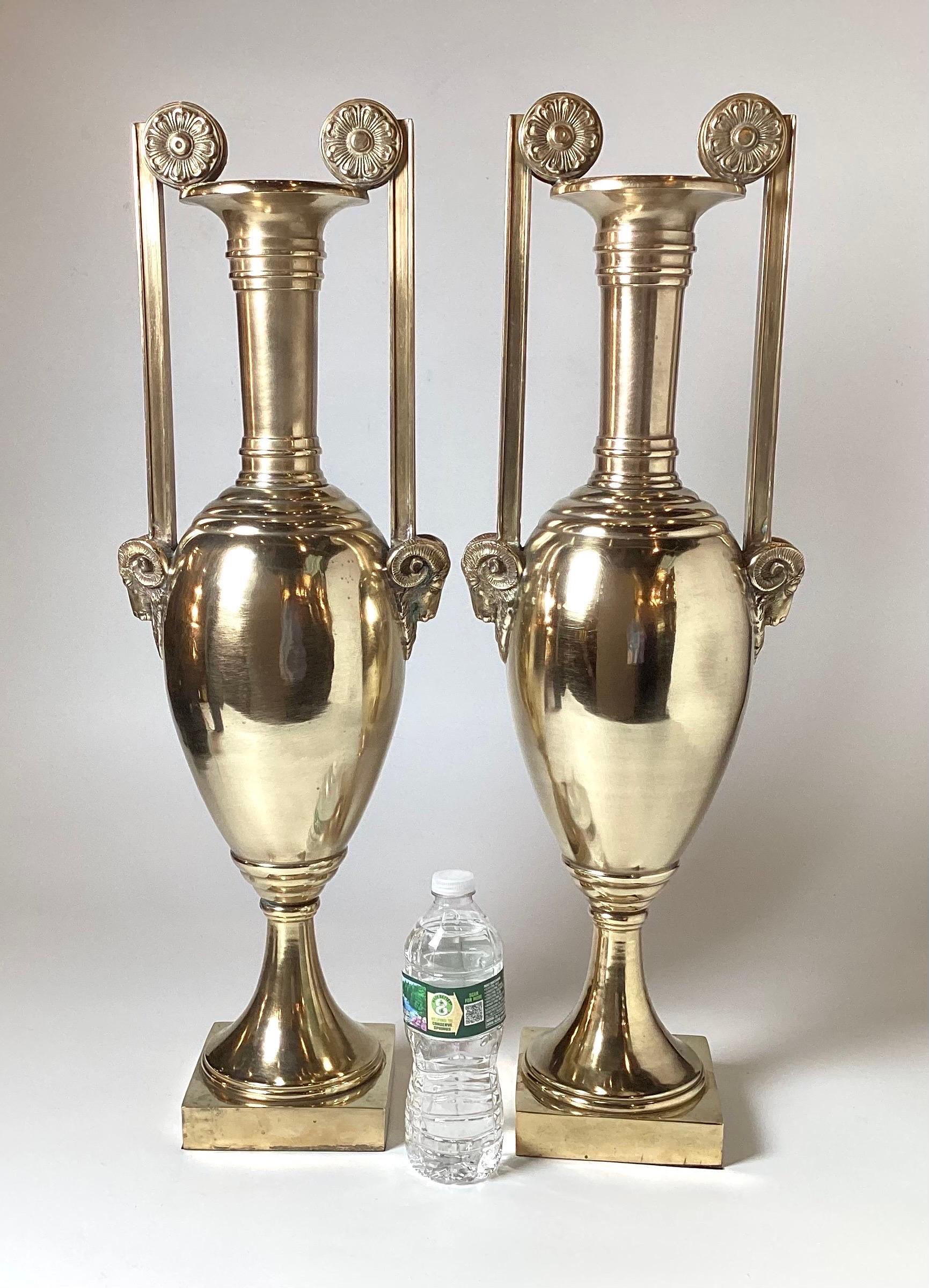 A handsome pair of elongated neoclassical cast and polished brass urns. The straight slender handles with cast rosettes at the tops and rams head detail at the bottom. The urns with a bulbous center resting on a 5.5 inch square pedestal base. Each