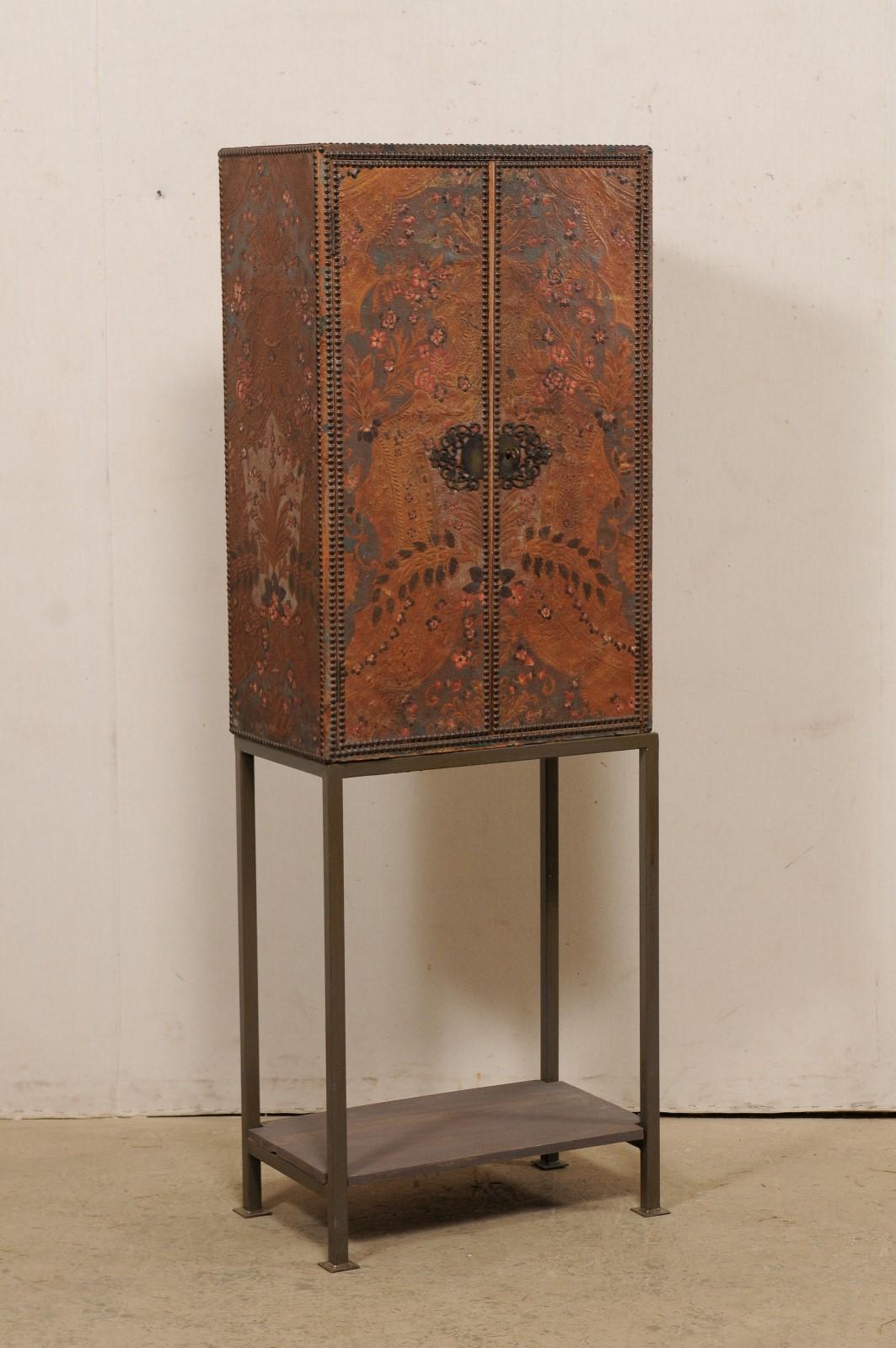 A Spanish embossed leather cabinet from the early 20th century, on newer custom iron base. This antique cabinet from Spain is beautifully wrapped in embossed leather with a floral motif, with nail head trim accents, and ornate hardware. The cabinet