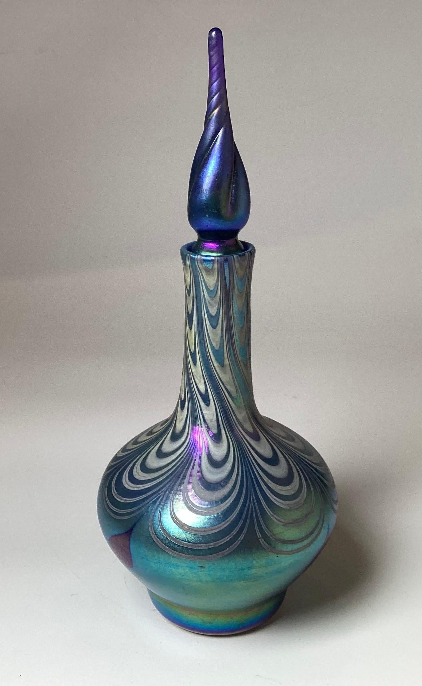 A tall Vandermark pull feather decanter. This piece is an intricately designed art glass bottle with a long twisted stopper. The body features a mesmerizing, swirling pattern in iridescent blues, greensand silvers creating an elegant and