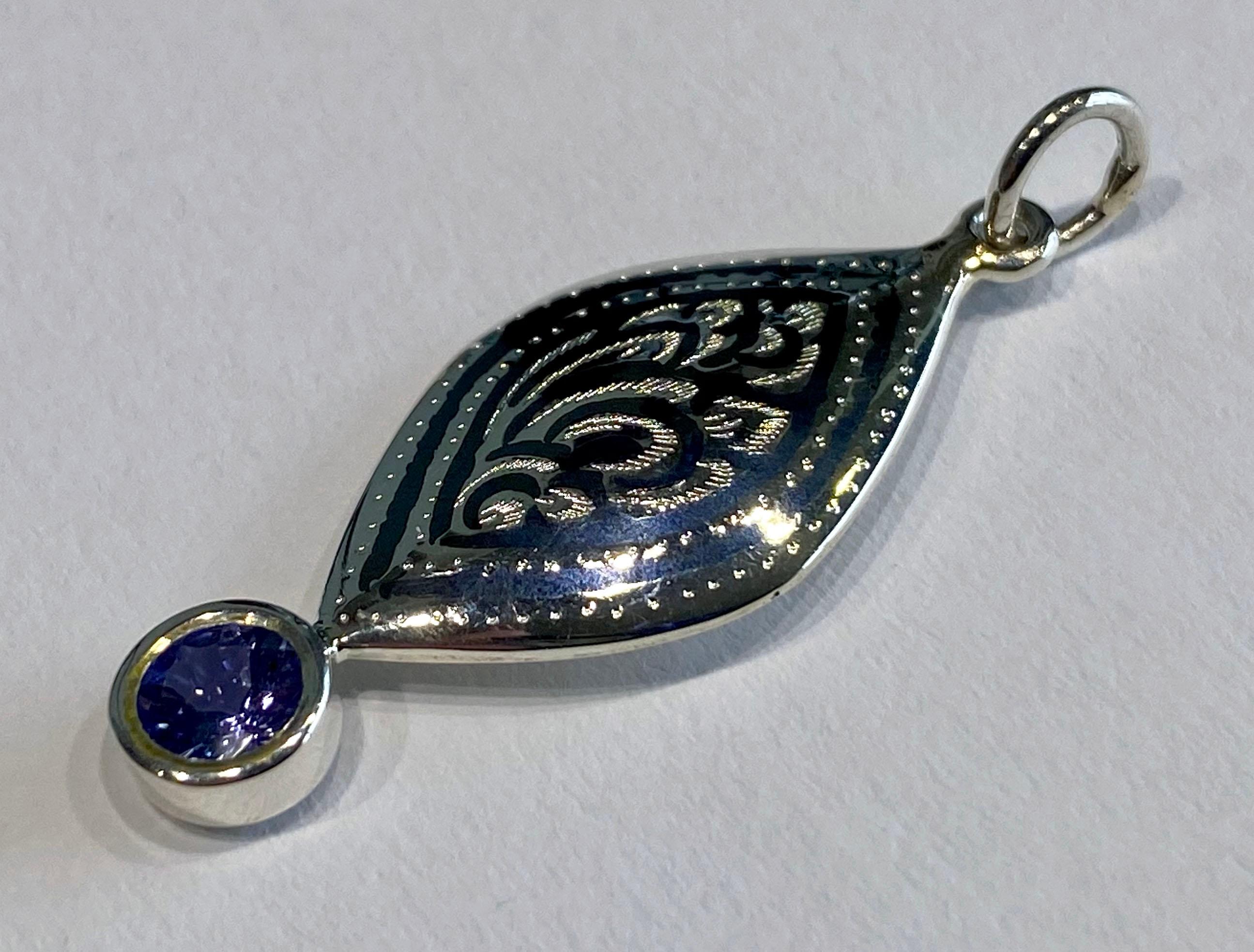 A Silver Filagree Pendant set with a Tanzanite. The style of Silver craft is called Nielloware that comes from Southern Thailand. This is a one of a kind pendant accented with a 5 MM Round Blue Tanzanite. The pendant hangs from an 18 inch silver