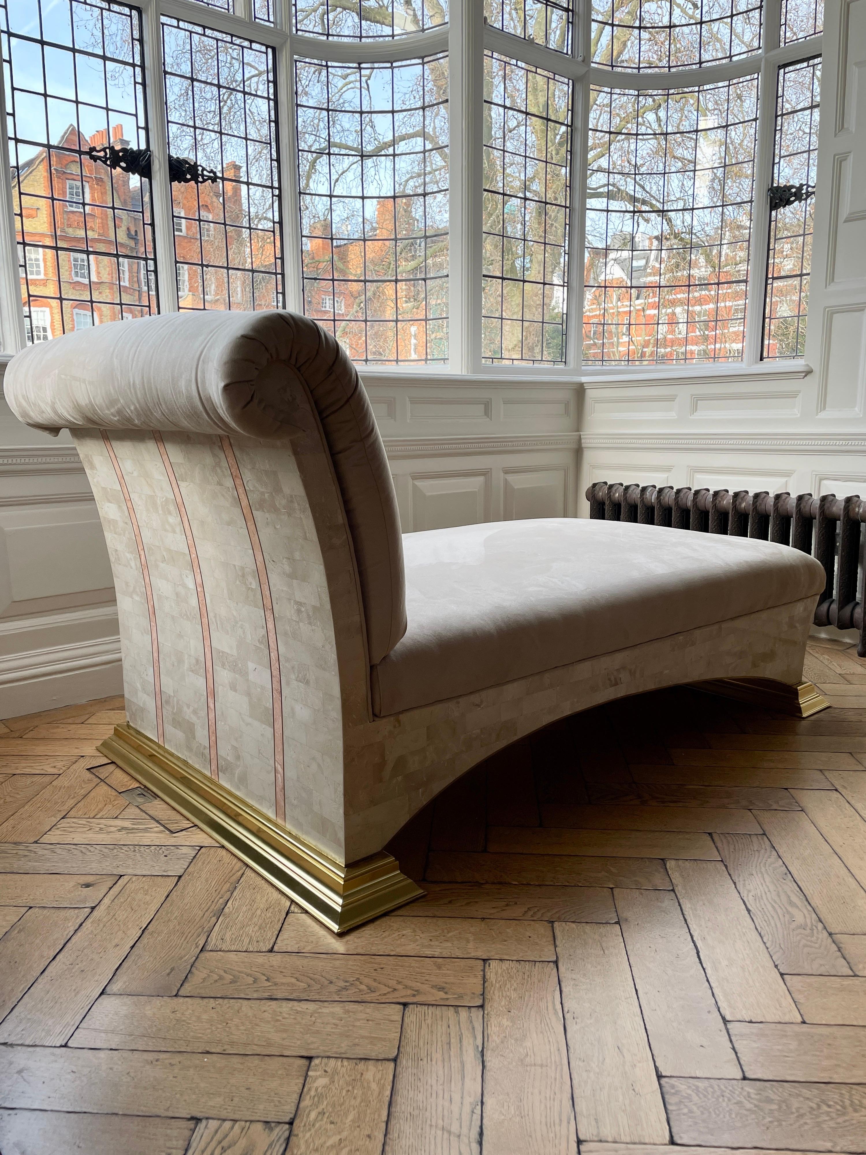 Un gran sofá cama con respaldo enrollado retapizado en ante sintético. La base arqueada se apoya en grandes zócalos escalonados de latón, de mármol teselado en colores naturales con decoración de mármol rosa en incrustaciones de latón. Una pieza