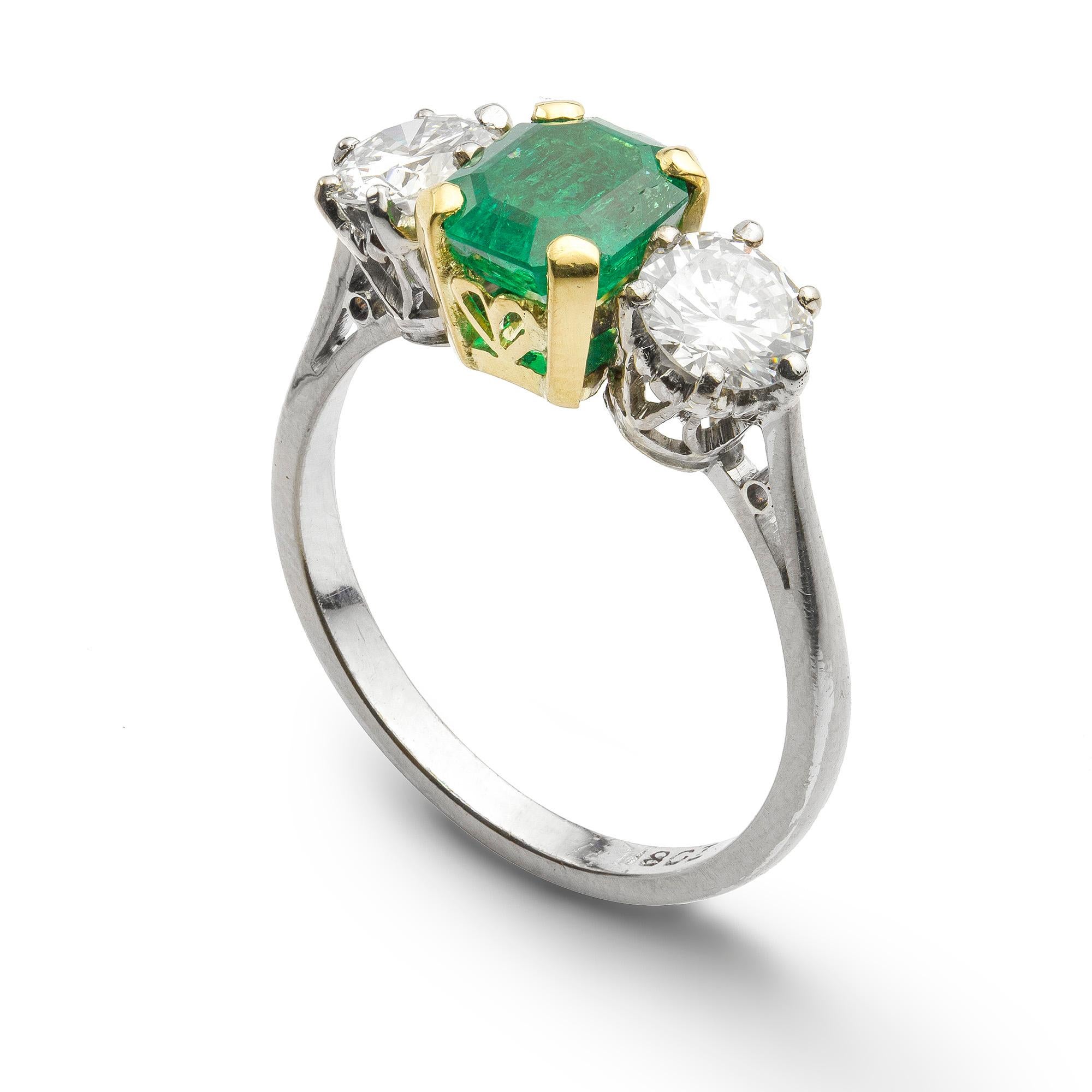 A three stone emerald and diamond ring, the central emerald-cut emerald, measuring approximately 7.0 x 6.2 x 4.2mm and weighing 1.34 carats, four claw set in gold, flanked by round brilliant-cut diamonds either side, weighing a total of 1.03 carats,