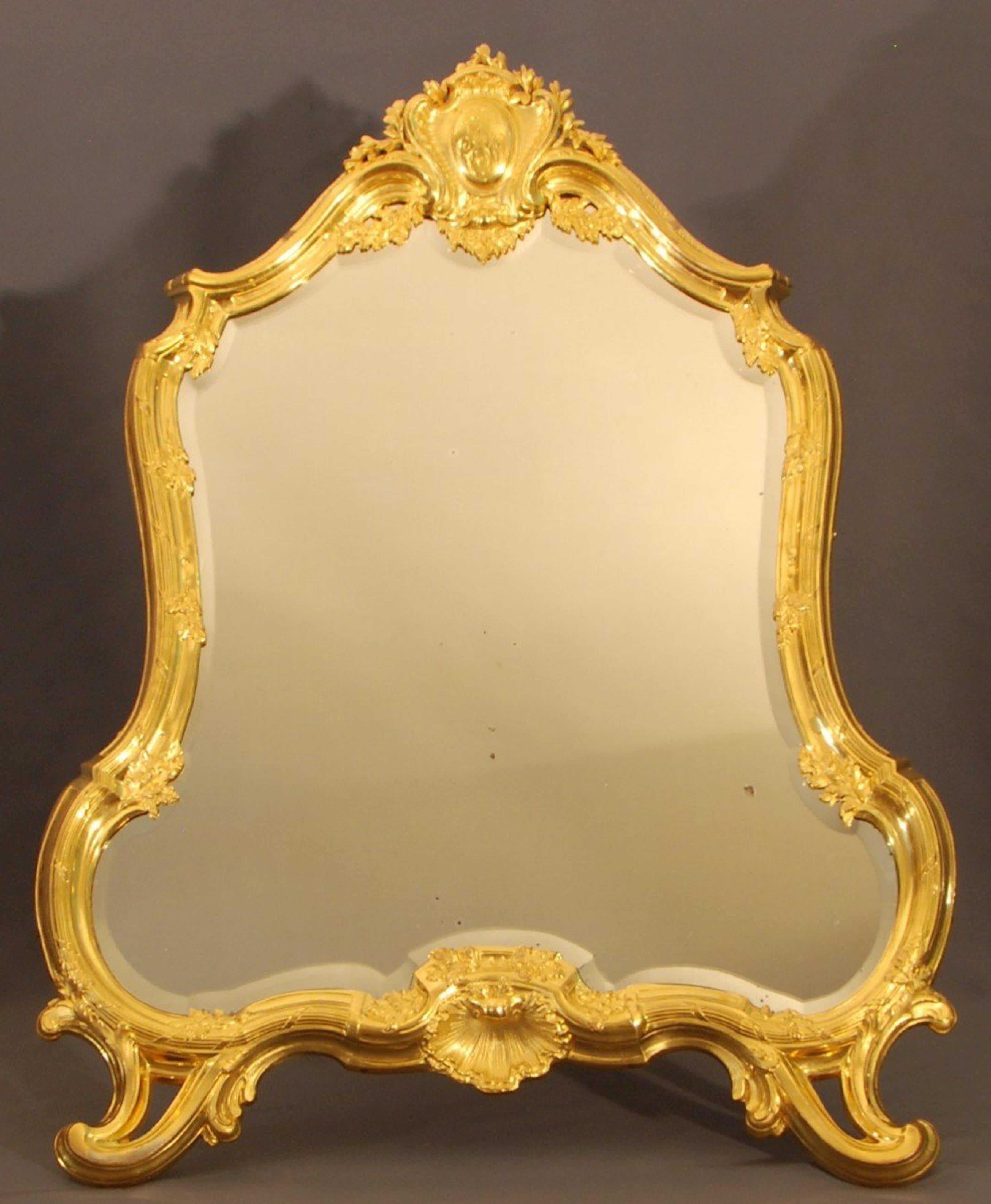 A toilet mirror in chiseled and gilded bronze, the bevelled mirror is surrounded by a moulding adorned with foliage scrolls. 
The feet in volutes frame a shell, with damping a coat of arms engraved with intials. 
On the back, a bronze square adjusts