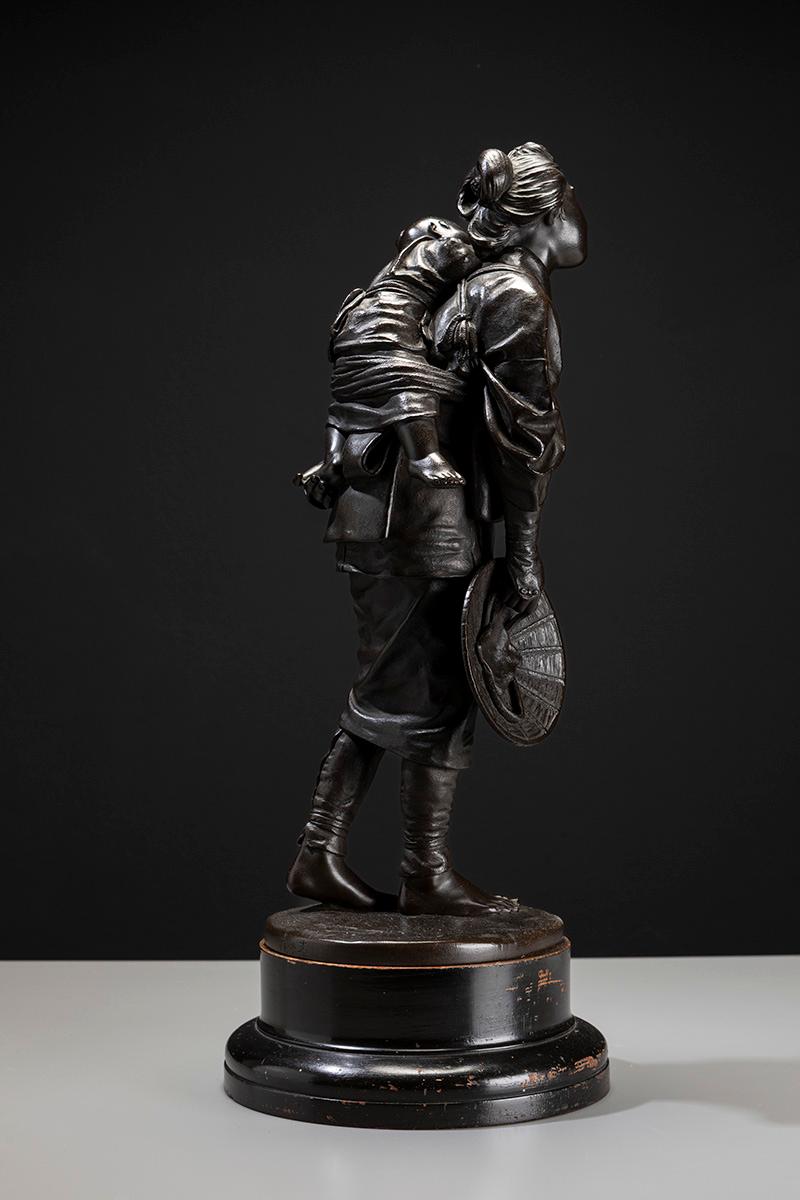 A Japanese bronze sculpture of the Tokyo School, depicting a young mother walking with her child tied to her back.
The woman wears a soft kimono fastened at the waist with an obi. Her torso leans slightly forward to support the child, who reaches