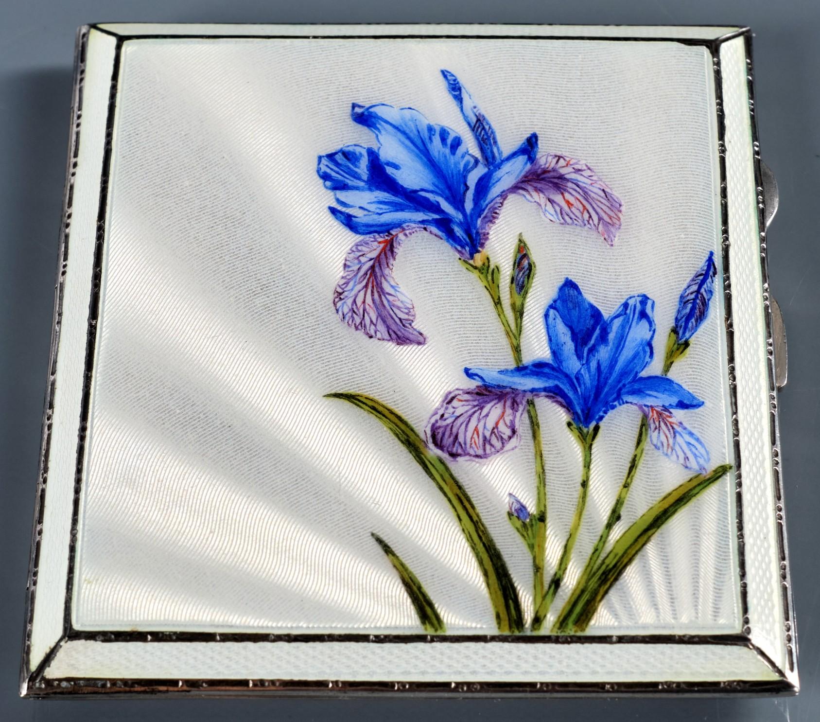 Die Vorderseite des Gehäuses zeigt eine exquisite handgemalte Tafel mit Irisblüten in Lavendel-, Königsblau- und Grüntönen vor einem strahlend weiß emaillierten, maschinell bearbeiteten Feld. Der Scharnierdeckel öffnet sich zu einem reich