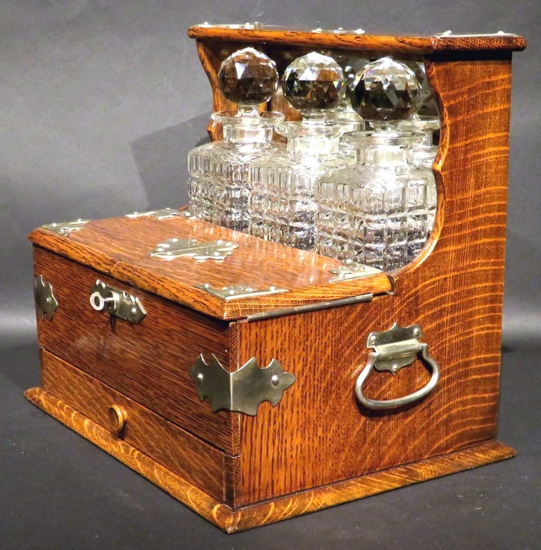 A very good early 20th century three bottle Tantalus in oak, showing a finely figured oak case with silver plated carrying handles & corner mounts, housing a mirrored & velvet lined interior fitted with three cut glass stoppered decanters fronted by