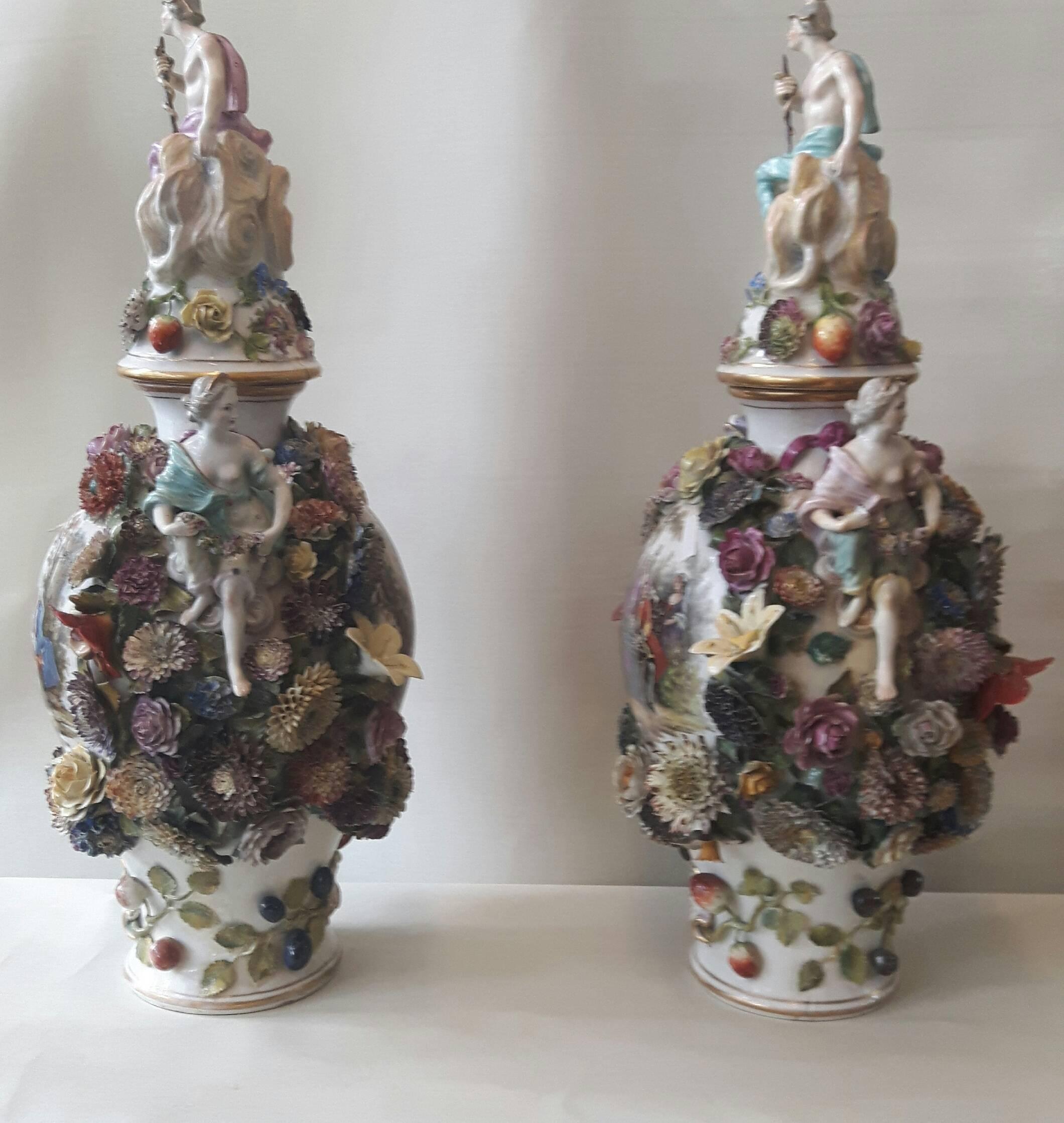 This pair of Dresden or Helena flower encrusted vases are in Meissen style.
The vases have hand painted cartouches after the French court artist Watteau and are adorned with hand-modelled encrusted flowers .On top of the domed lids sit a pair of