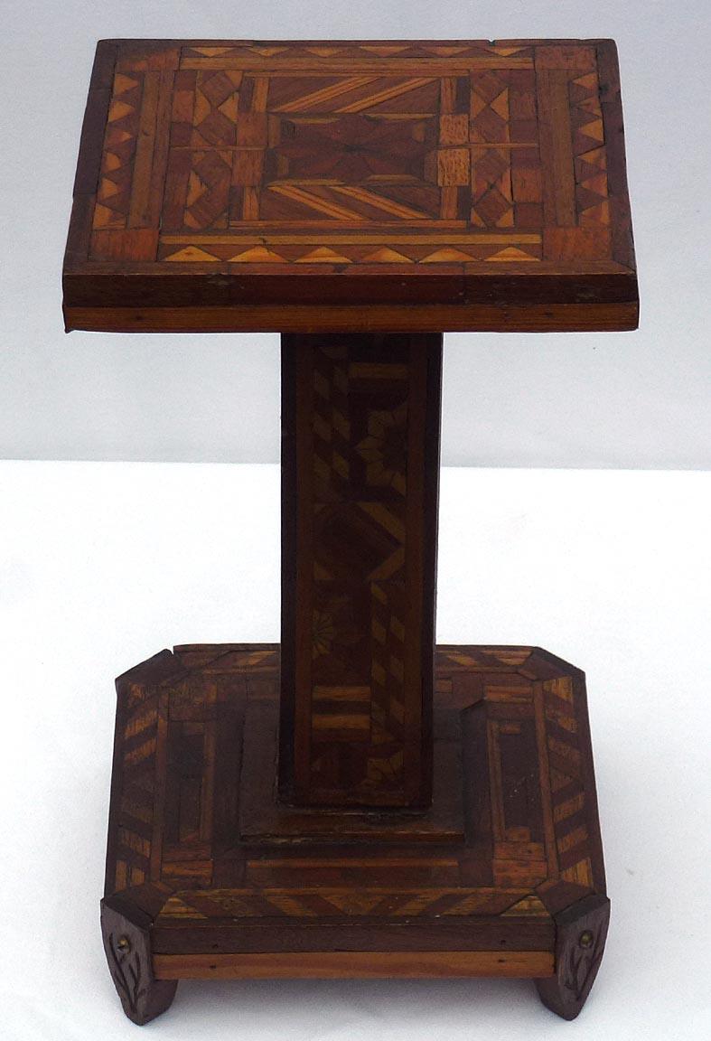 This is a very intricate small marquetry pedestal stand. Each side of the upright is decorated in a different pattern. The four feet also have incised designs. The different colors of wood are finished in a clear lacquer or varnish. It stands 13