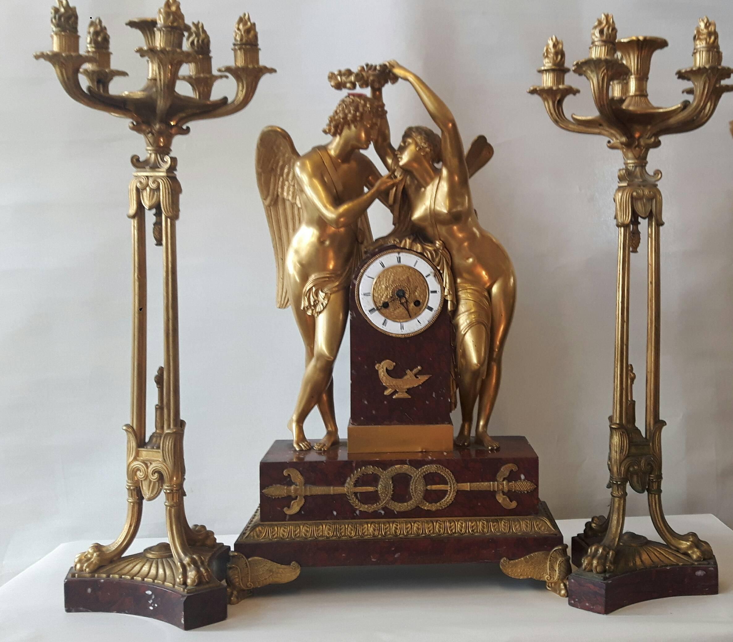 A wonderful garniture in marble and ormolu, the central clock face engraved in ormolu and surrounded by enameled numerals. The clock is adorned by two amorous ormolu figures of an angel and a nymph and the body is decorated with Roman designs in the