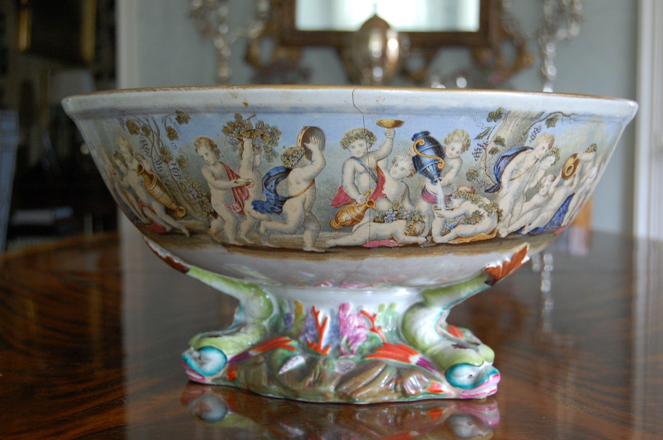 The transfer decorated pottery bowl with its interior decorated with five drunken Bacchic putti beneath a frieze of other frolicking and drunken Bacchic putti, the similarly decorated exterior raised on an elaborate sea life base moulded with three