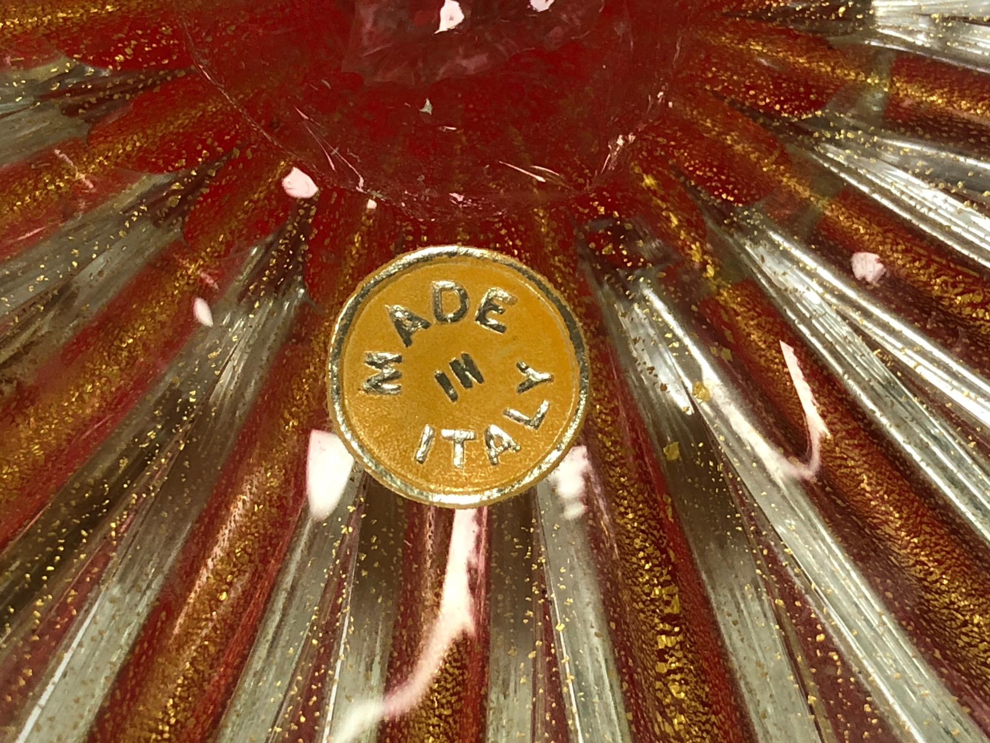 El cuenco lobulado, de un vivo rojo fuego, se alza sobre un pie claro, todo con inclusiones de metal dorado (aventurina), con etiqueta original de papel de aluminio.