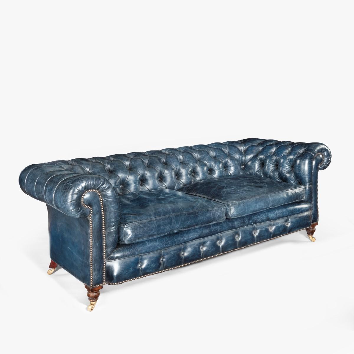 A Victorian 2-seater leather Chesterfield sofa, of typical form with rolled over arms and back, set on turned walnut front legs with the original castors and out-swept back legs, re-upholstered in deep-buttoned distressed blue leather. English,