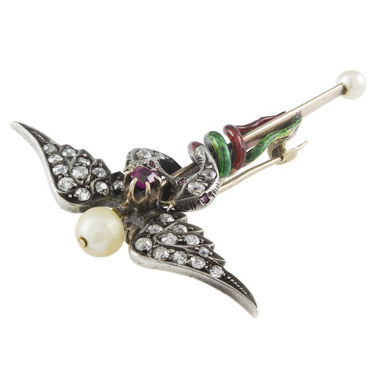 A Victorian caduceus brooch, a pair of old brilliant-cut diamond-set snake heads set with ruby eyes and a green and a red enamelled tail surmounted by a pair of old brilliant-cut diamond set wings and a ruby centre to a gold staff terminated with