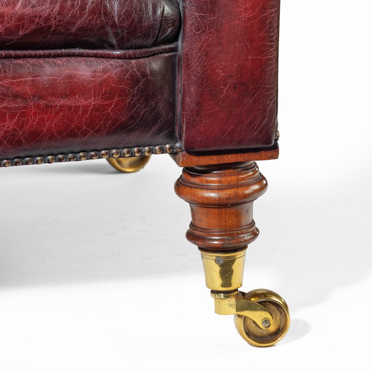 A Victorian deep buttoned Chesterfield settee, with an upright back and scroll arms, set on four turned walnut legs with brass castors, reupholstered in distressed burgundy leather. English, circa 1850.

 