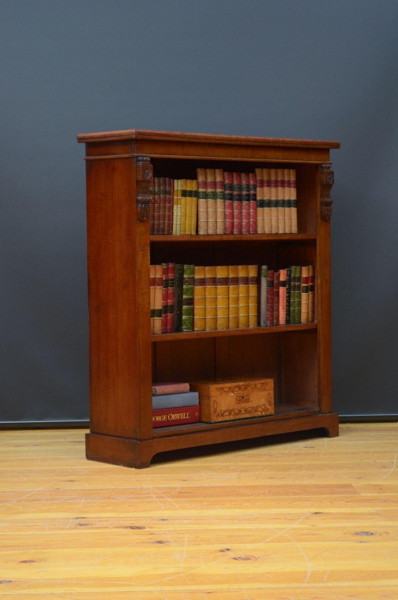 K0301 Victorian open bookcase in figured mahogany, having figured mahogany sides and figured mahogany top with moulded edge above two height adjustable shelves flanked by drop carvings, all standing on shaped plinth base. This antique bookcase is in