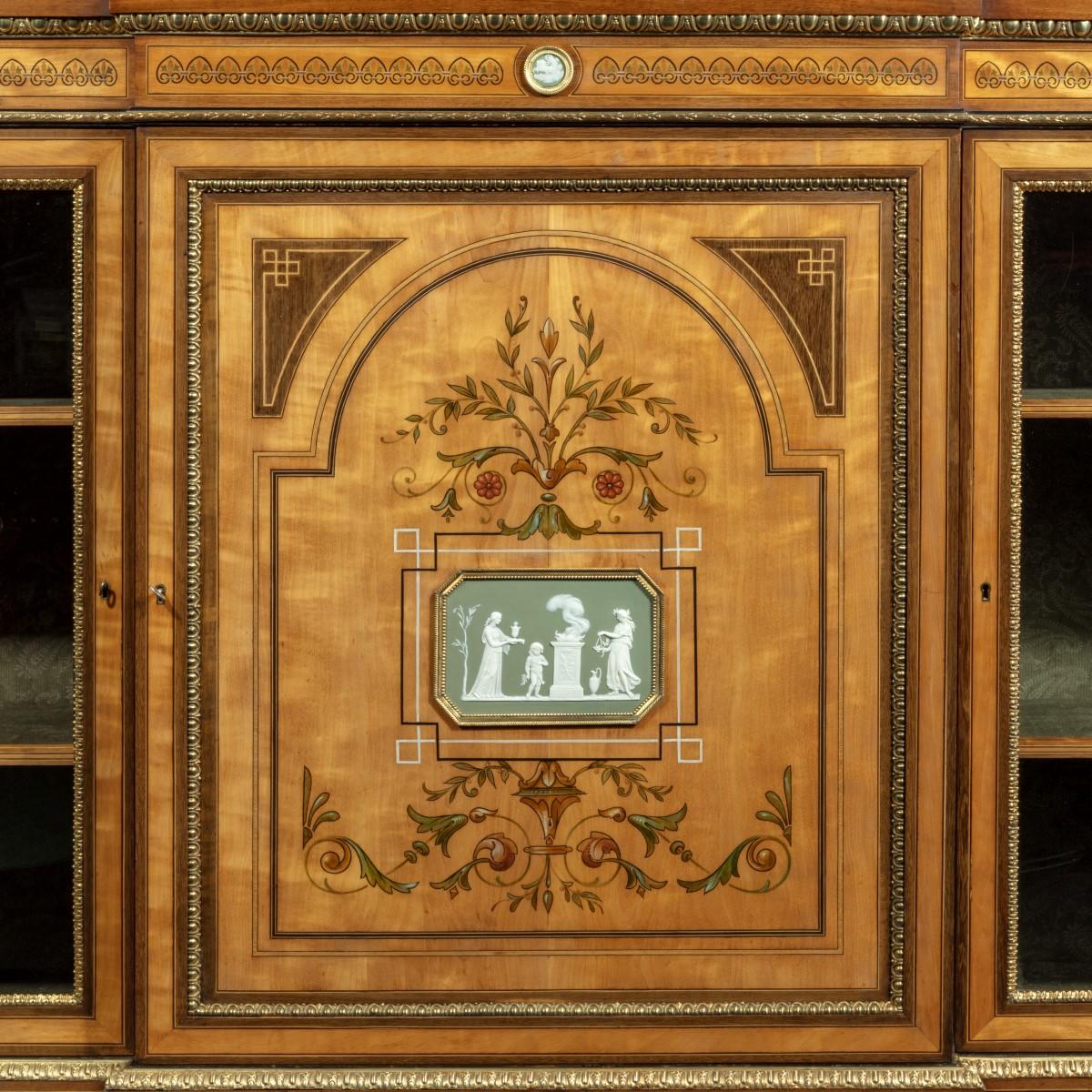 A Victorian satinwood breakfront side cabinet with Wedgwood plaques attributed to Dyer and Watts, the shaped top above a central door flanked by two glazed doors enclosing shelves, decorated with stencilled floral motifs, two pale green Wedgwood