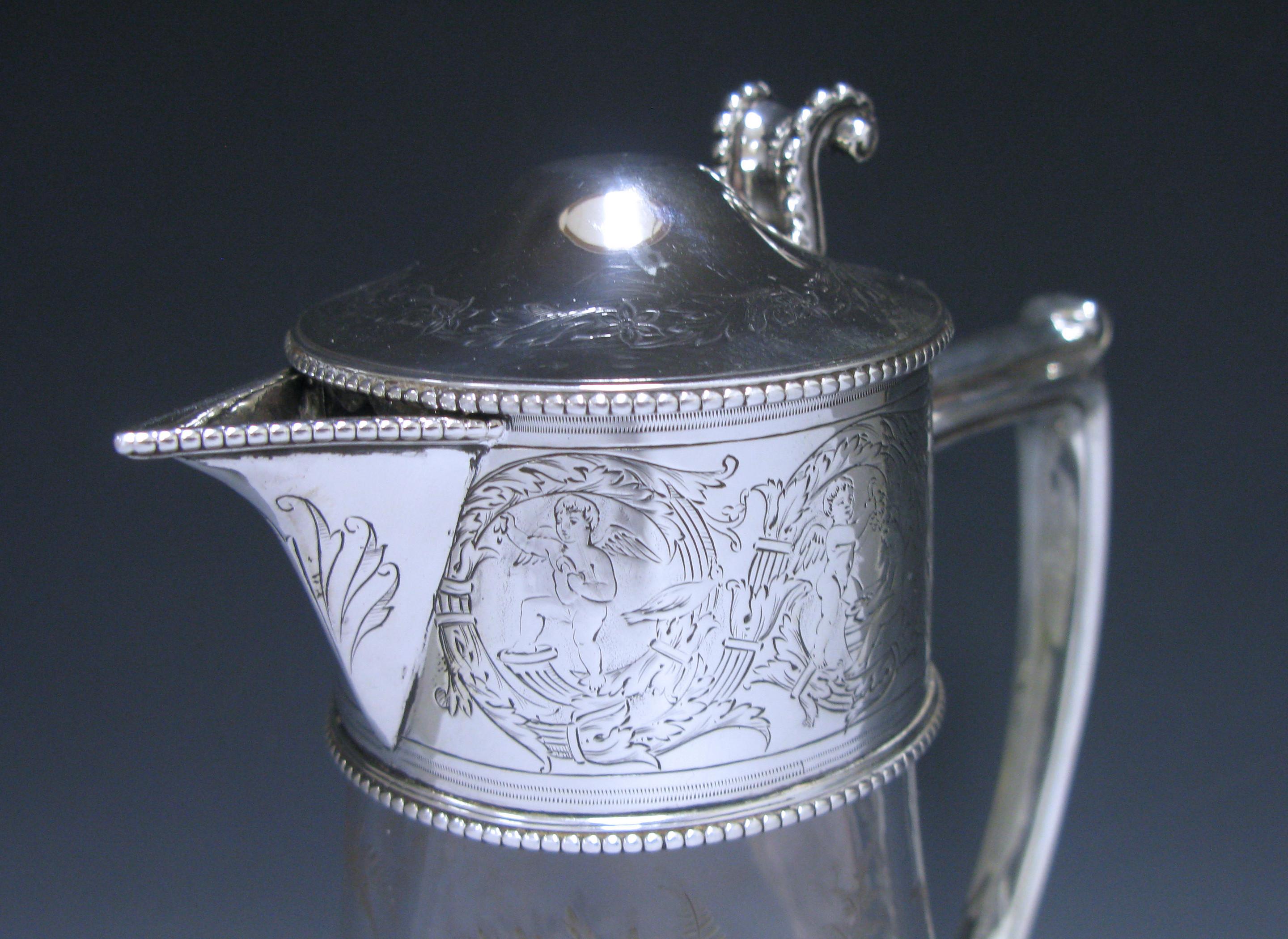 A splendid Victorian silver mounted Claret Jug. The hinged mount is engraved with cherubs on one side and on the other is central plain cartouche joined by swags of leaves and flowers. The domed lid has a border of leaves. The beautiful glass body