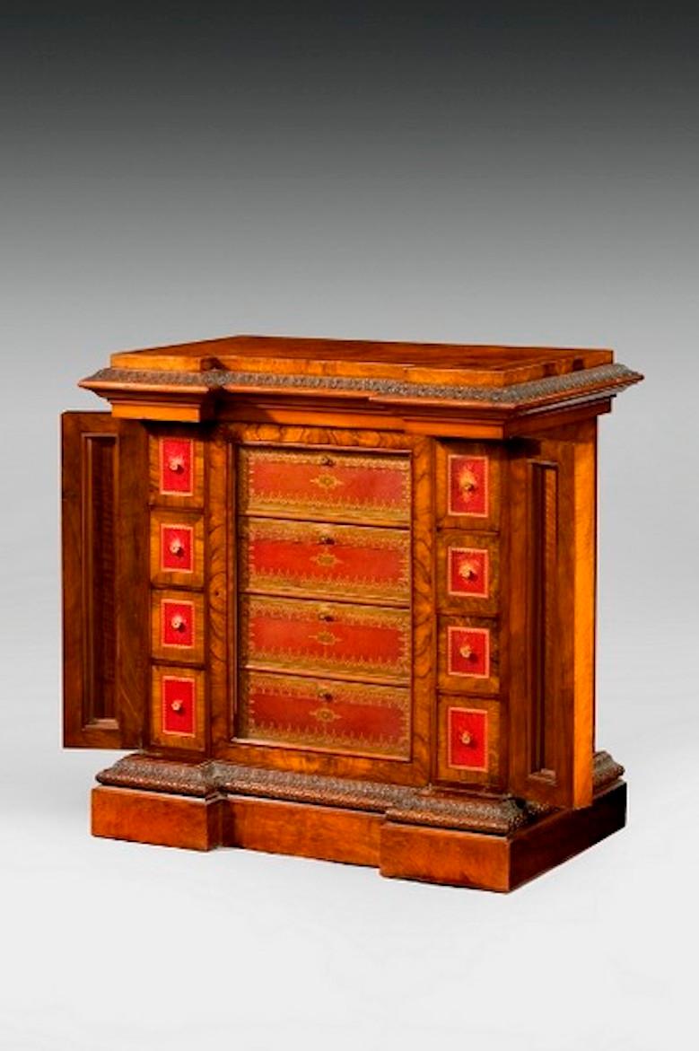 This walnut cabinet has a shaped, stepped top above a central glazed door enclosing four Moroccan leather drawers with cedar linings. Flanked by eight smaller disguised drawers, the ornately carved corner pilasters open to reveal further small