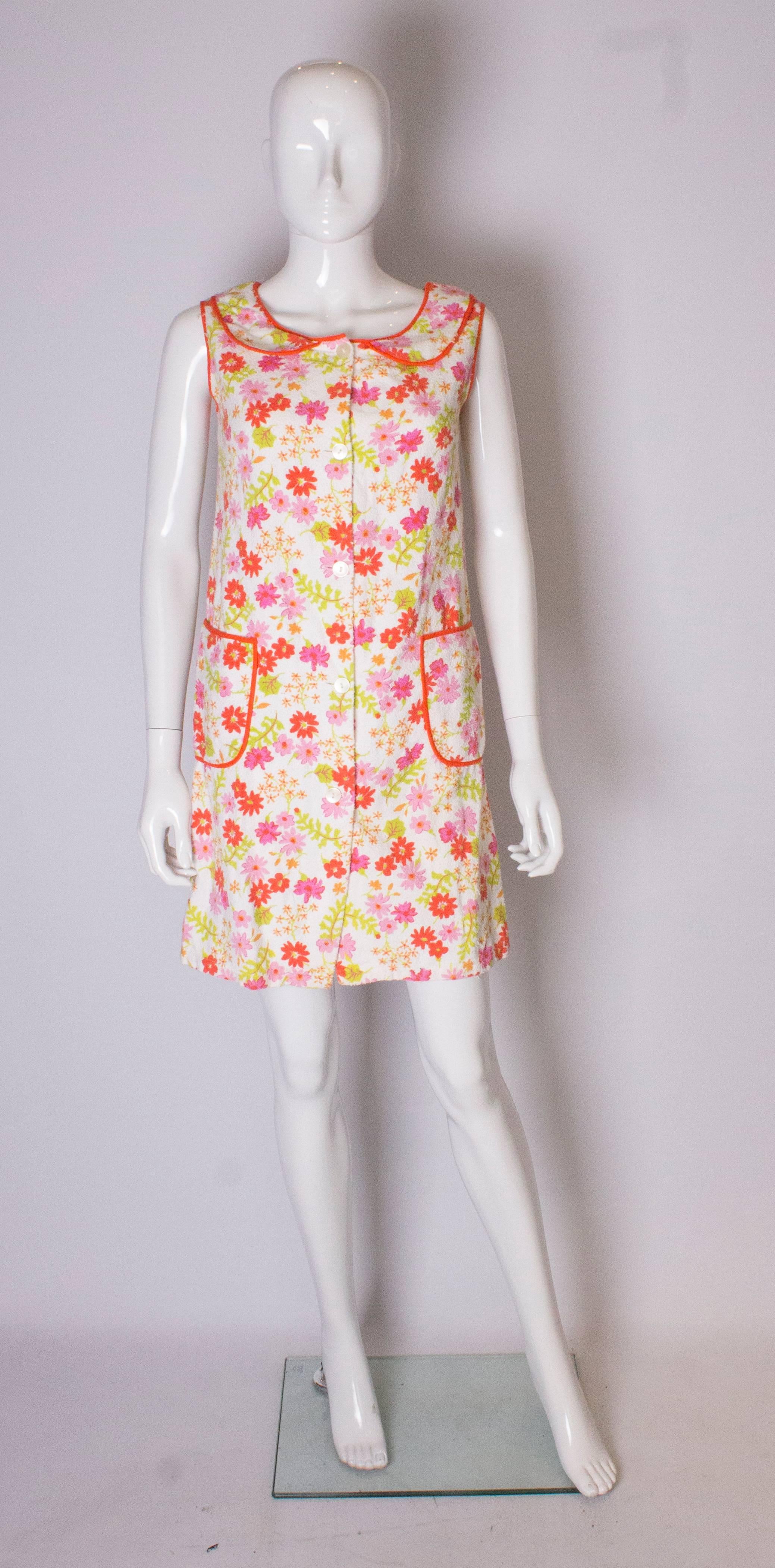 A pretty cotton towelling  summer dress for city or coast.   The dress has a white background with a pink, red and orange floral print. It has a peter pan collar,  two pockets and  a five button  front  opening.