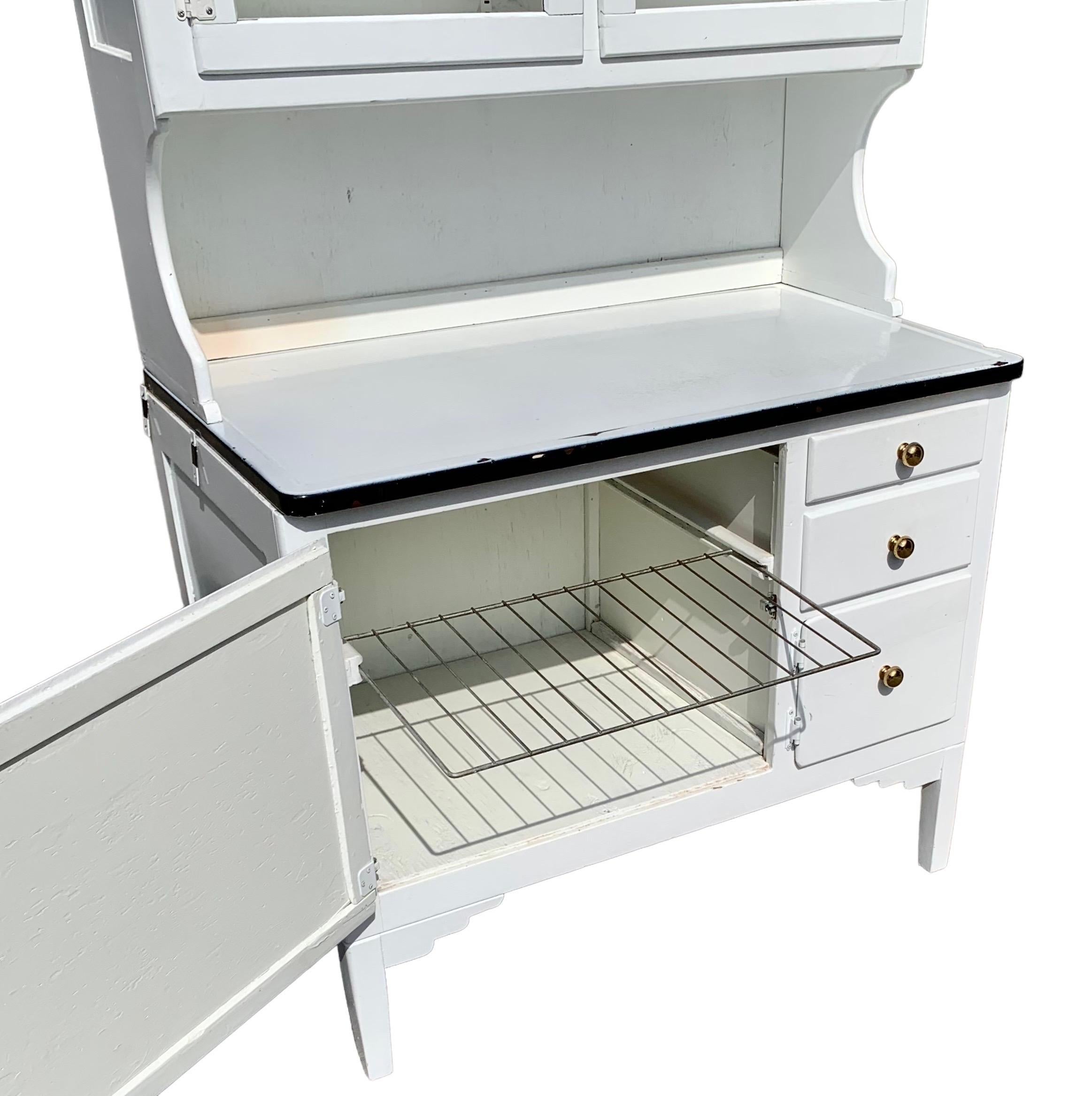 An American Art Deco, white painted hardwood and porcelain Hoosier cabinet having two glass doors and a shelf above a porcelain work surface, above a door with pull out, metal cooling grill, two drawers, and one small door. 

A very useful piece,