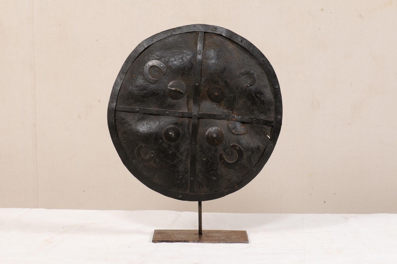 An Ethiopian tribal shield on custom iron stand. This vintage shield, originating from Ethiopia, has an overall convexly rounded-shape, and is made of a rich brown animal hide with wonderful patina. The leather shield is decorated with tooled metal