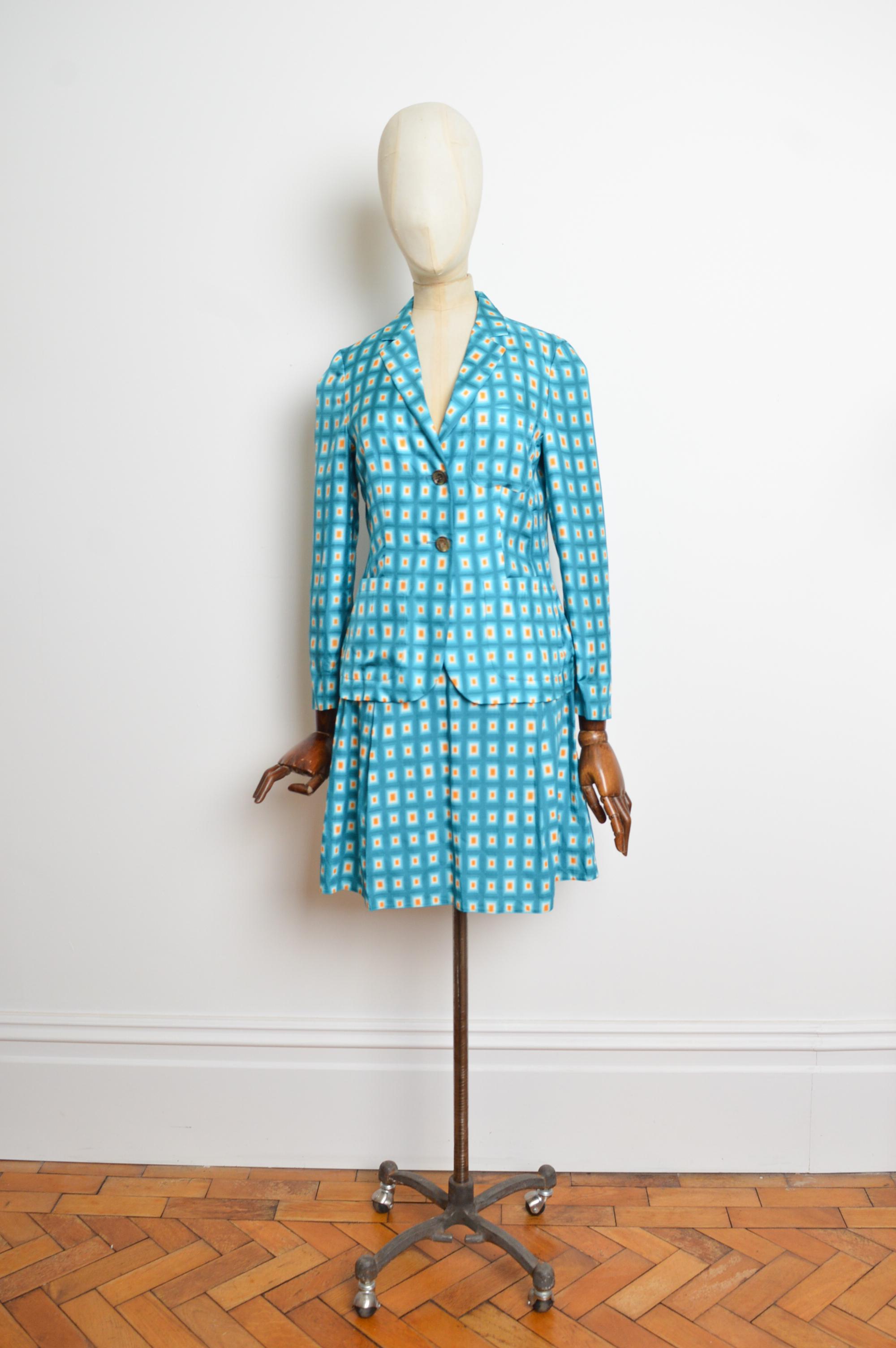 Superba tuta con stampa geometrica di
Nella collezione autunno/inverno 2012-2013 di Miuccia Prada, i colori sono caratterizzati da vibranti tonalità di blu e giallo. 
Il blazer presenta 2 bottoni sul davanti e la gonna a pieghe ha una chiusura