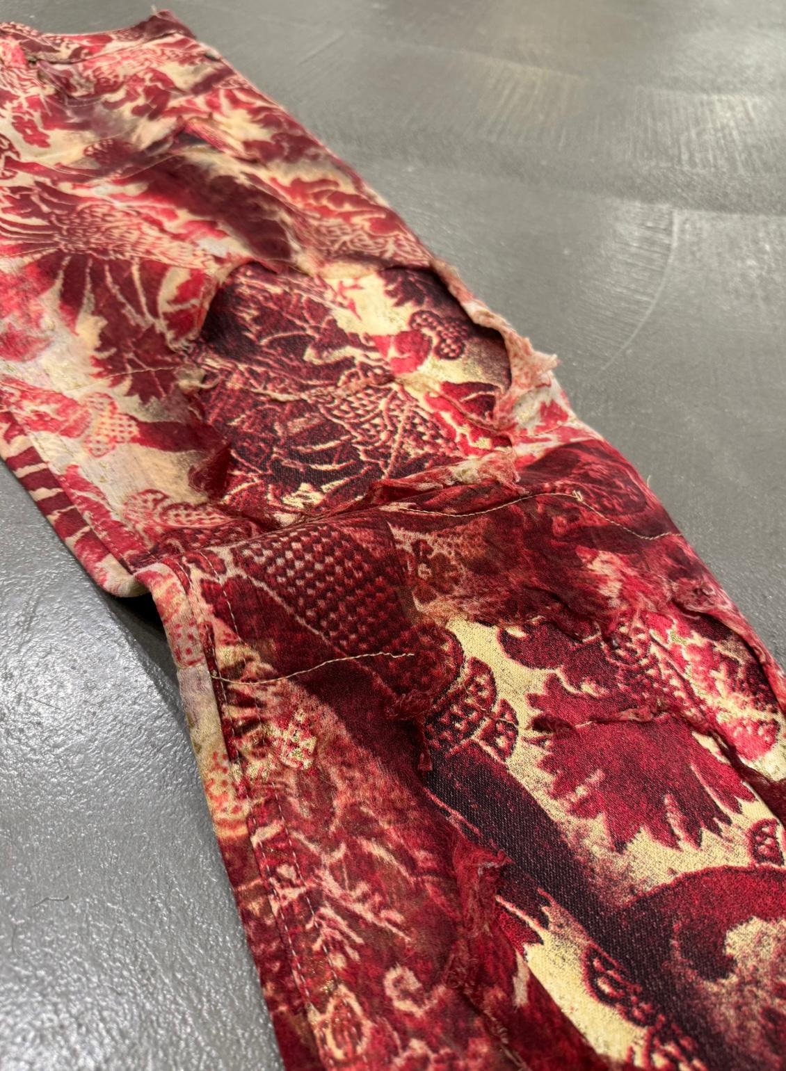 These Roberto Cavalli runway pants from the Autumn/Winter 2001 collection are a bold statement piece. Featuring a double-layered design, the pants showcase a distressed “burned” effect, blending rich red and gold tones in an intricate pattern. The