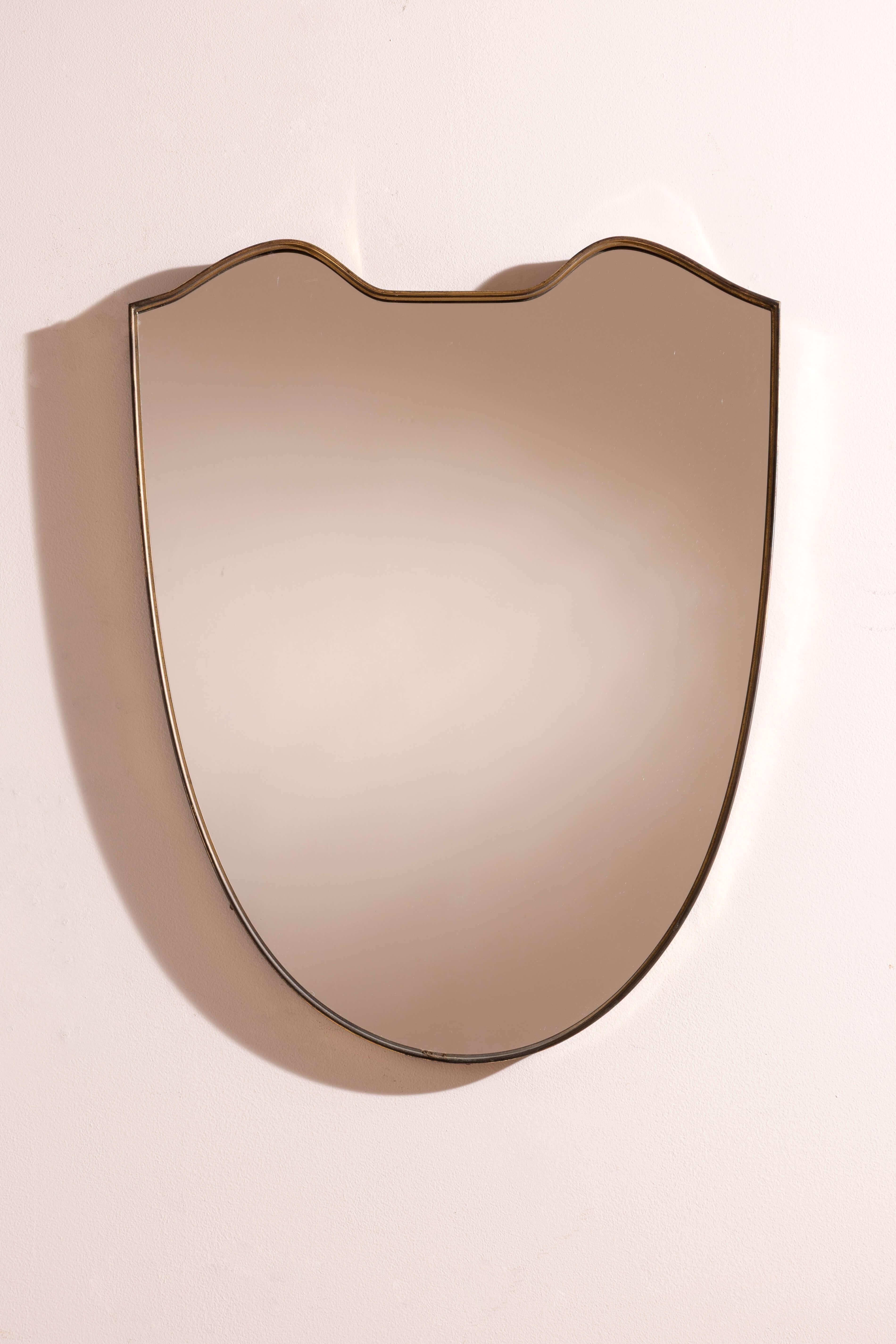 Miroir mural élégant avec cadre en laiton, Italie, années 1950.
La monture en laiton, élancée et délicatement moulée, suit discrètement les contours sinueux du bouclier, lui conférant à la fois raffinement et sobriété. La patine naturelle du métal,