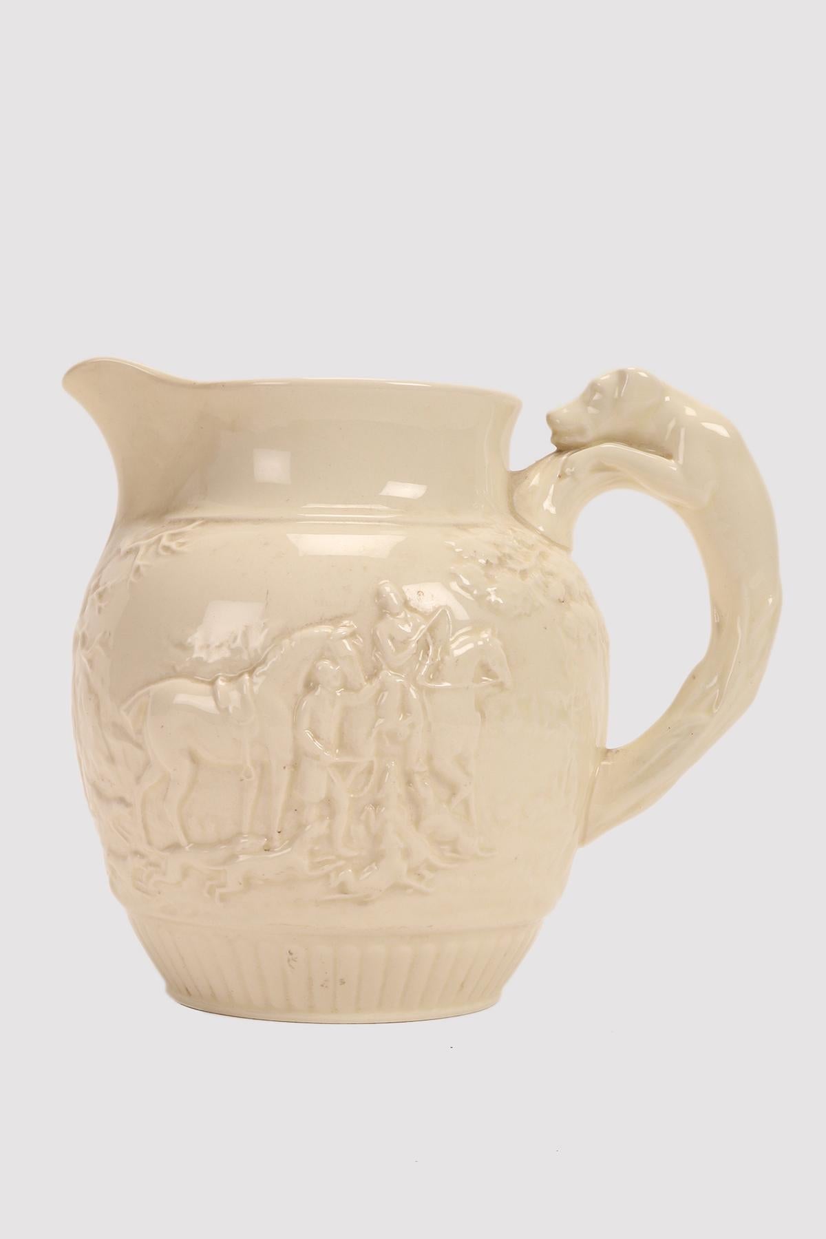 The jug is made of fine pottery with a cream colored glaze cover. The flared body shows elements in relief in the first elevation. Then a central band with a fox hunting scene with trees and a gate in low relief. The rim is distinct, flared, with an