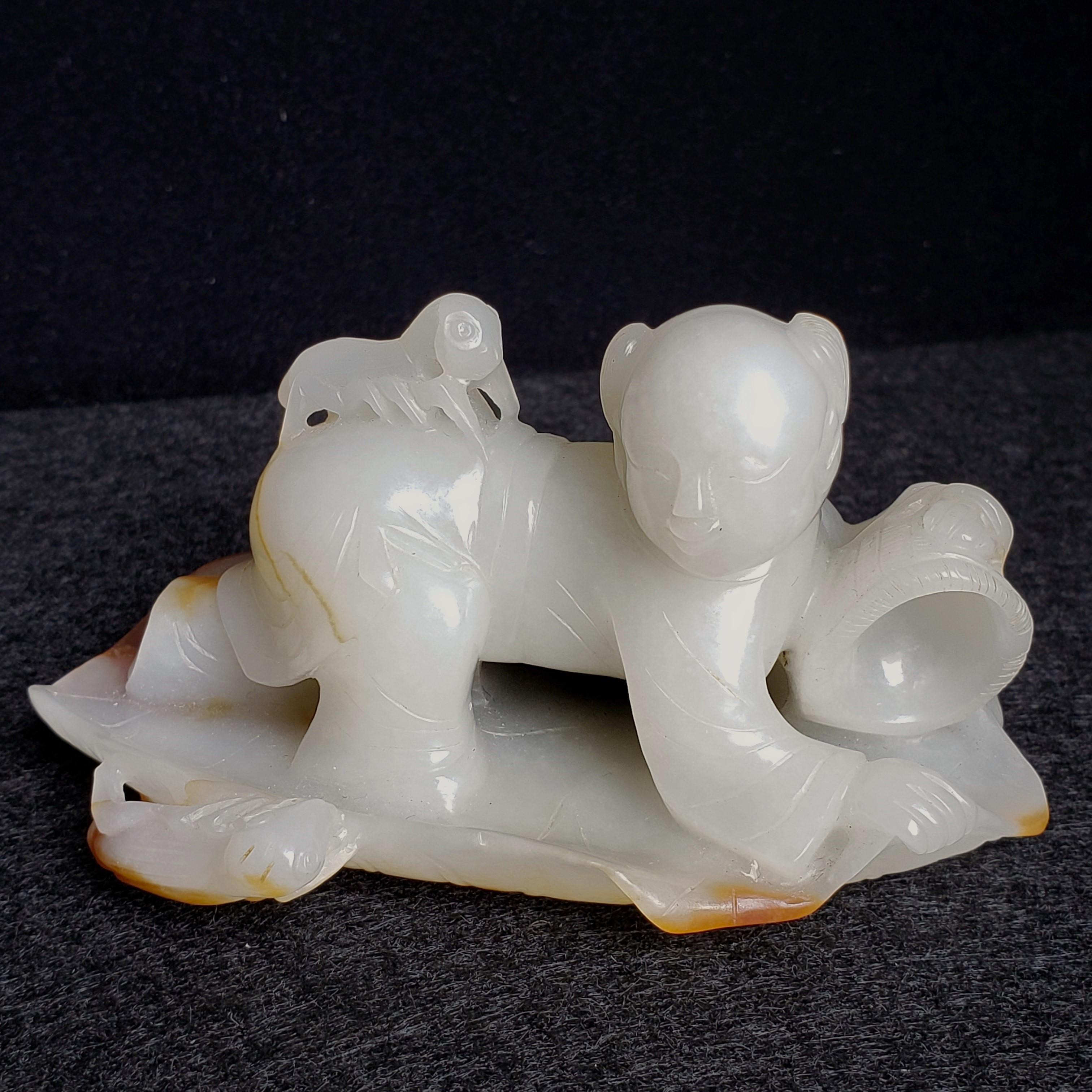 Habilement sculpté en jade de Hetian, ce garçon vêtu d'une longue robe est accroupi sur une feuille de lotus allongée montée par un grillon. Il saisit un panier de la main gauche tandis qu'un autre grillon se perche sur son dos. Le jade est d'une