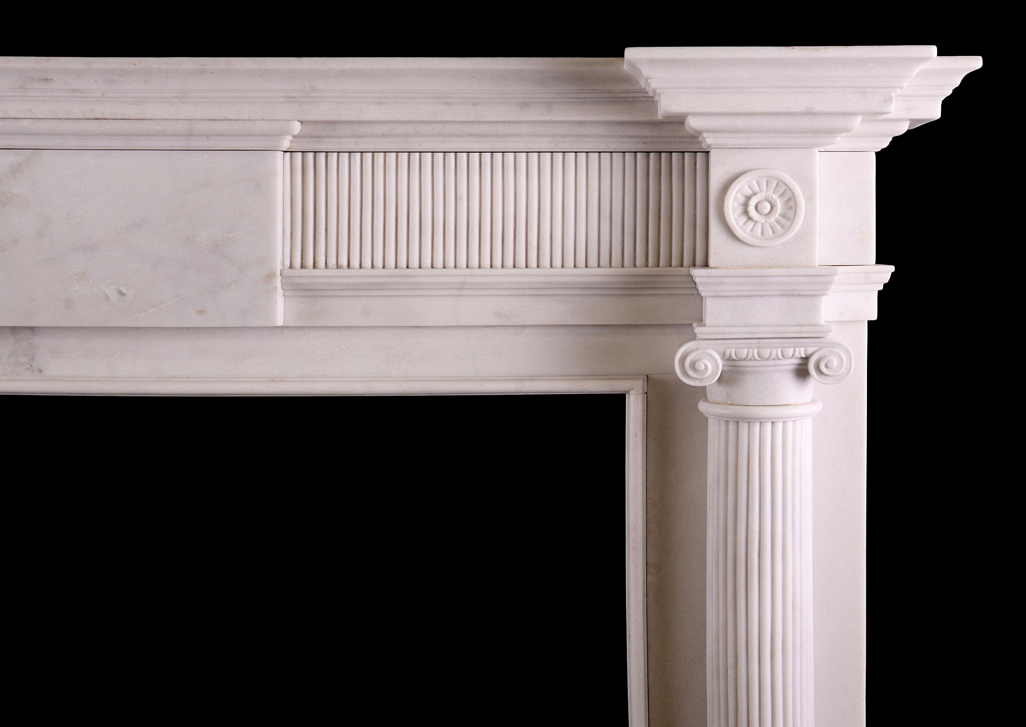 A white marble fireplace in the Georgian manner. The jambs with reeded, tapering columns, surmounted by Ionic capitals with egg and dart carving. The frieze with plain centre blocking, flanked by reeding and round rosette paterae. Moulded,