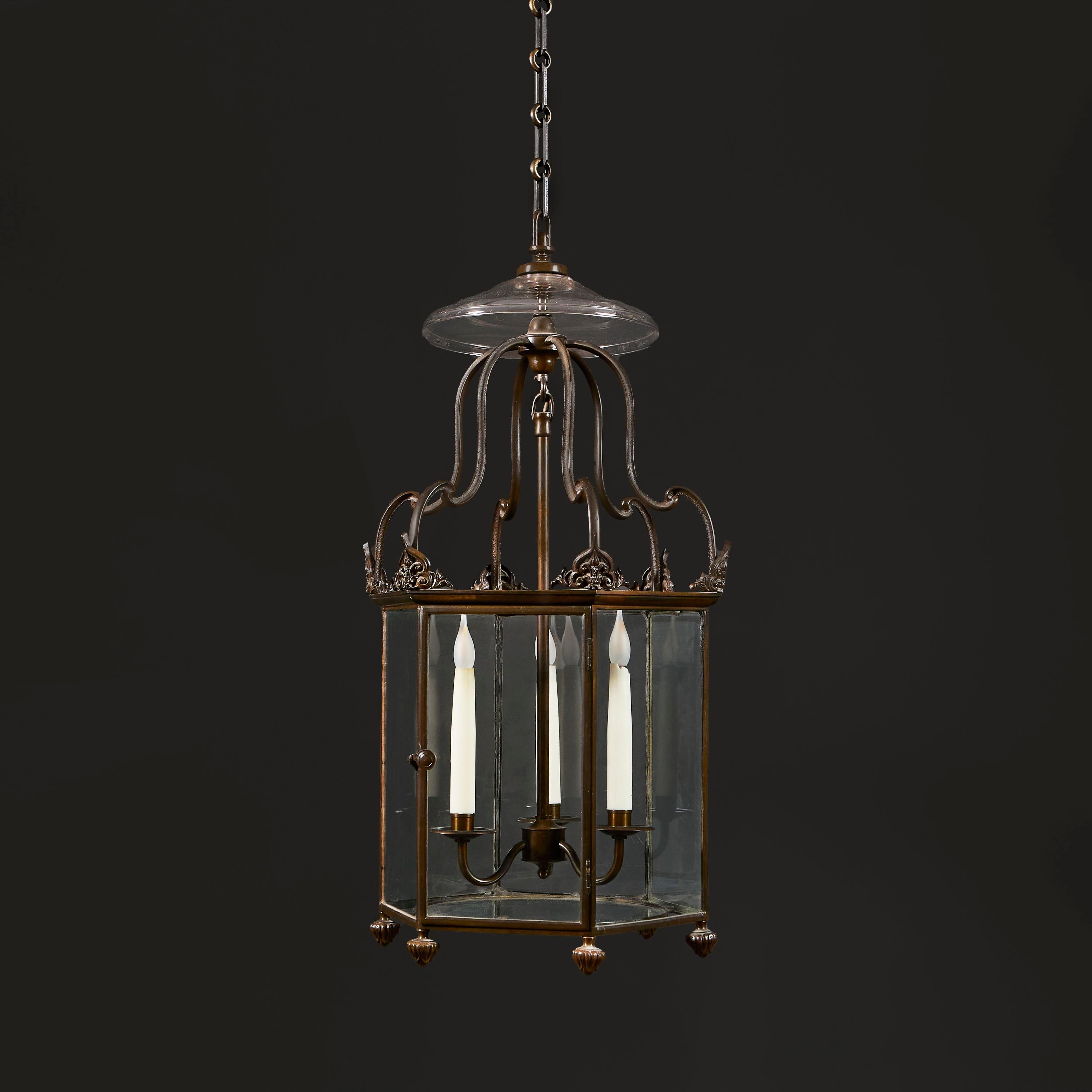 England, circa 1840

A fine early nineteenth century hexagonal bronze hall lantern with stylised anthemions to the canted corners, shaped uprights below a glass cowl, all supported on turned feet decorated in foliate design. One foot of chain